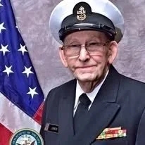Elderly man in military uniform standing in front of an American flag.