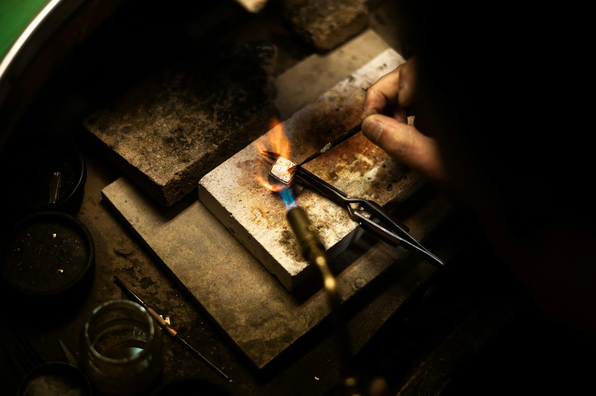 A person using a blowtorch to heat a small metallic object in a workshop or jewelry making setting, with tools and equipment surrounding the workspace.
