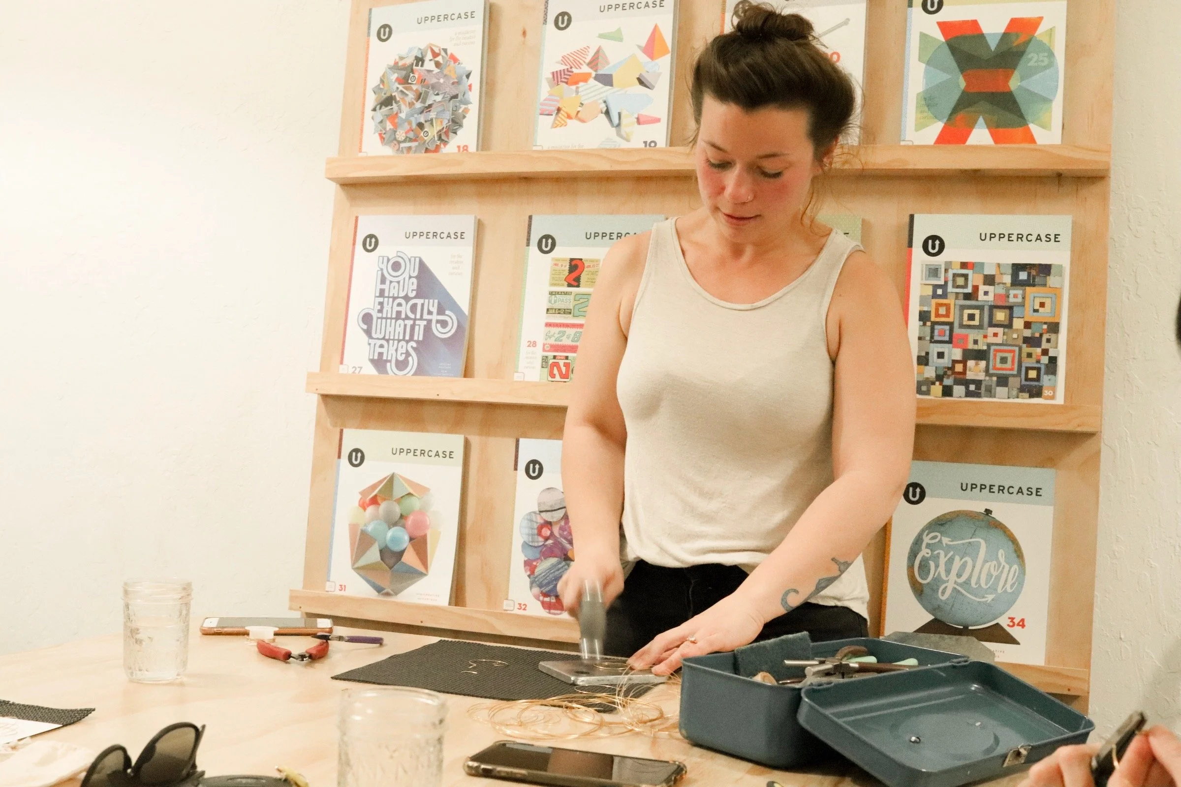 A woman with dark hair in a bun and tattoos on her arms is working on a craft project at a table, with various tools and supplies around her. Behind her, a wooden bookshelf displays colorful posters about geometric shapes and art projects.