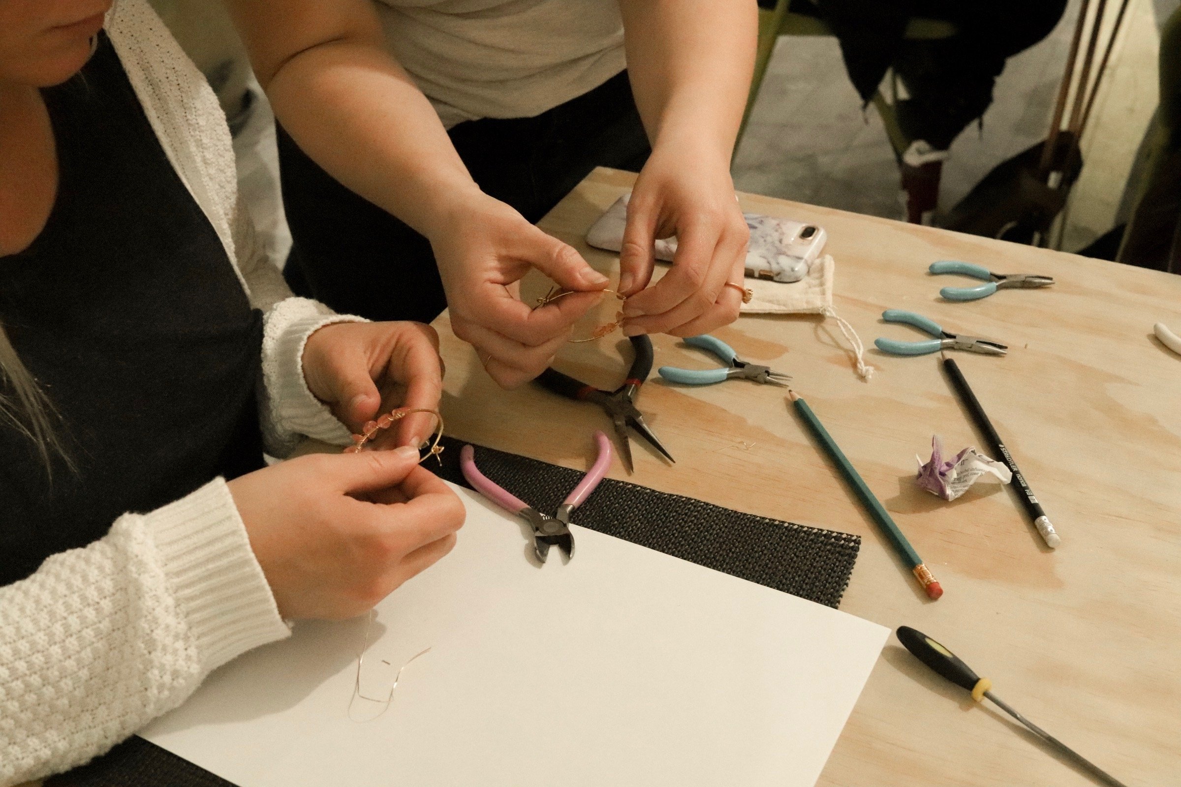 People working on jewelry crafts at a table with tools and materials, including pliers, wires, and a smartphone.