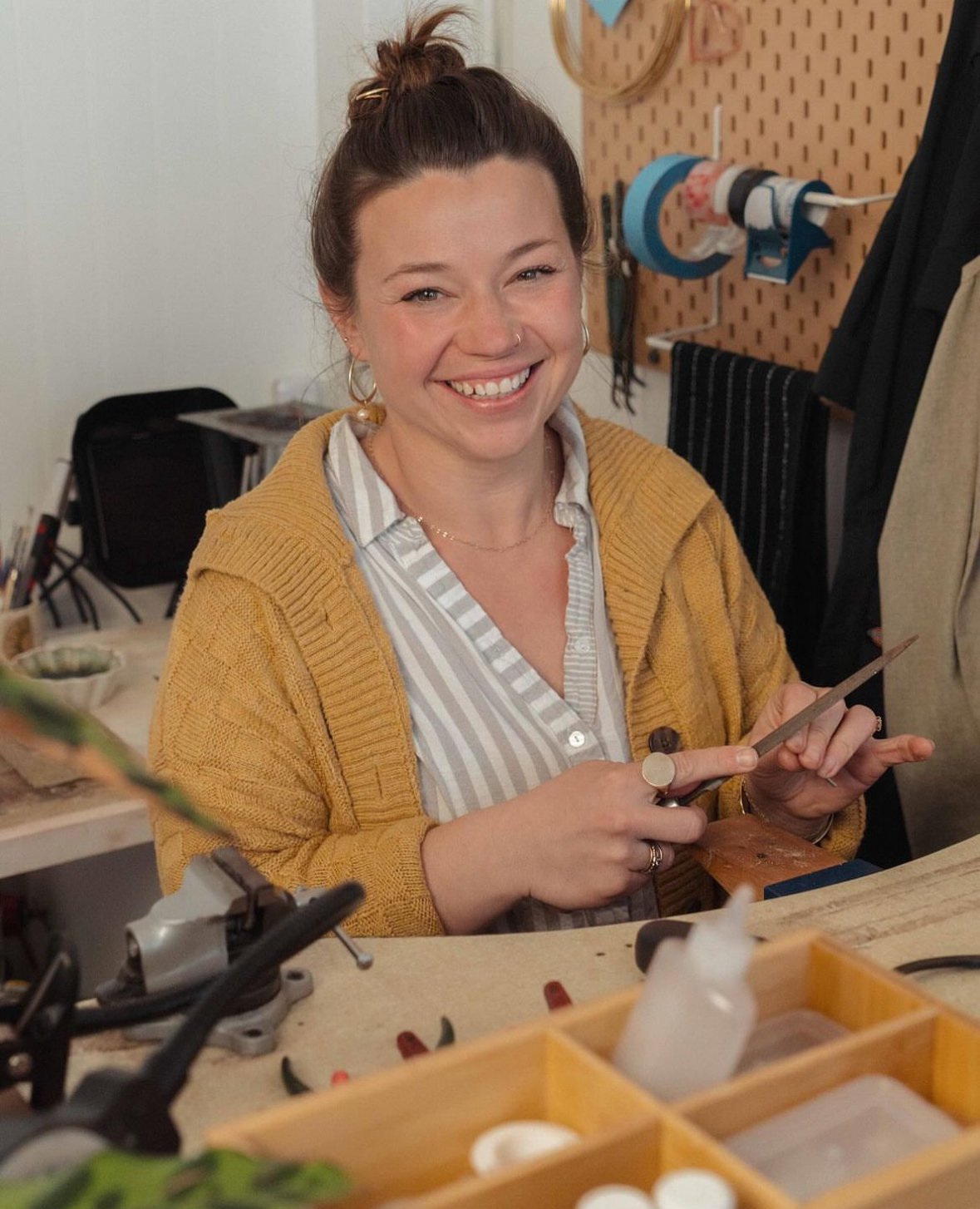 A smiling woman with a bun hairstyle, wearing a striped shirt and mustard yellow cardigan, is holding jewelry tools in a workshop with tools and materials in the background.