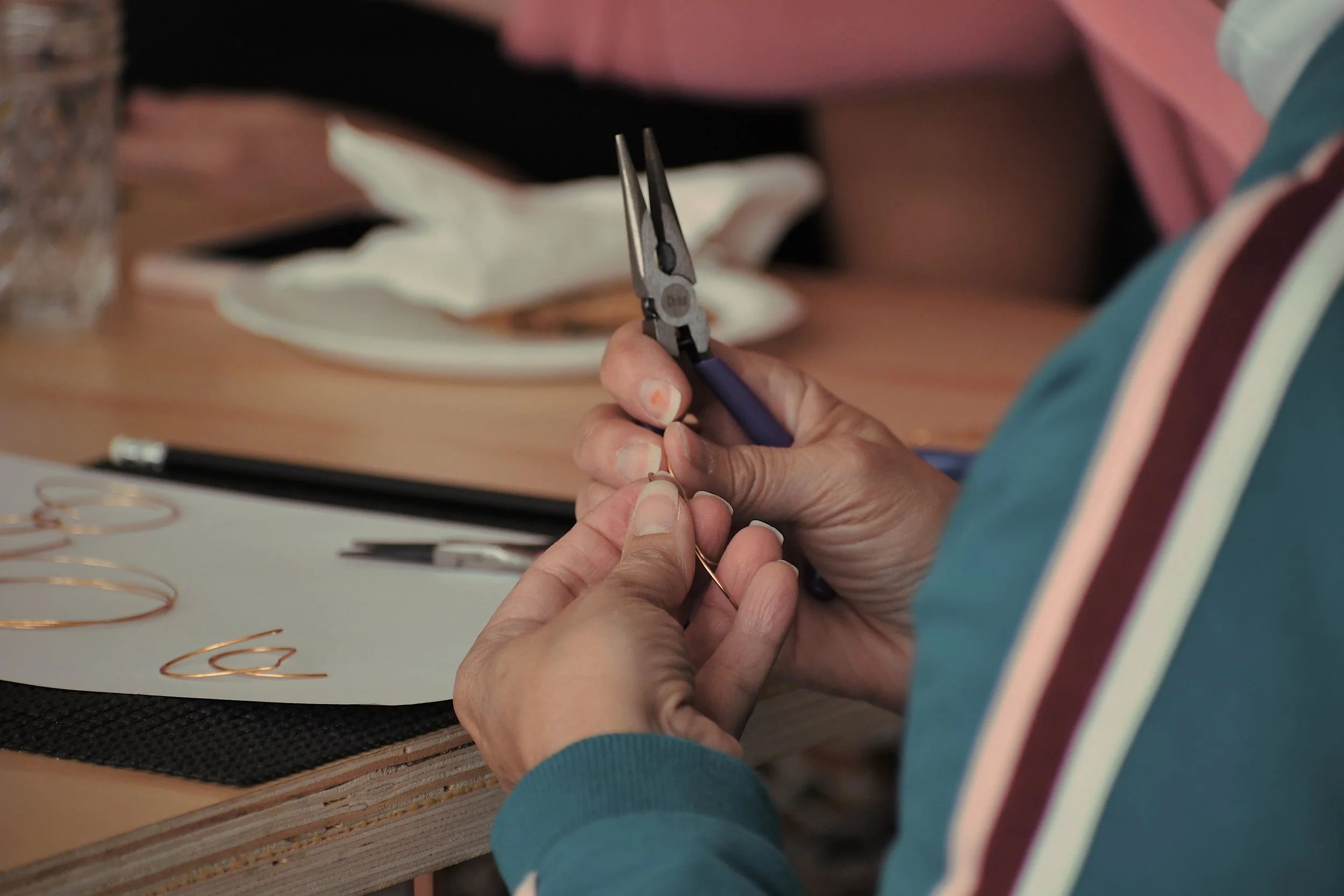 Person using pliers to manipulate a thin wire at a desk work area with gold-colored paper clips and a wooden surface.