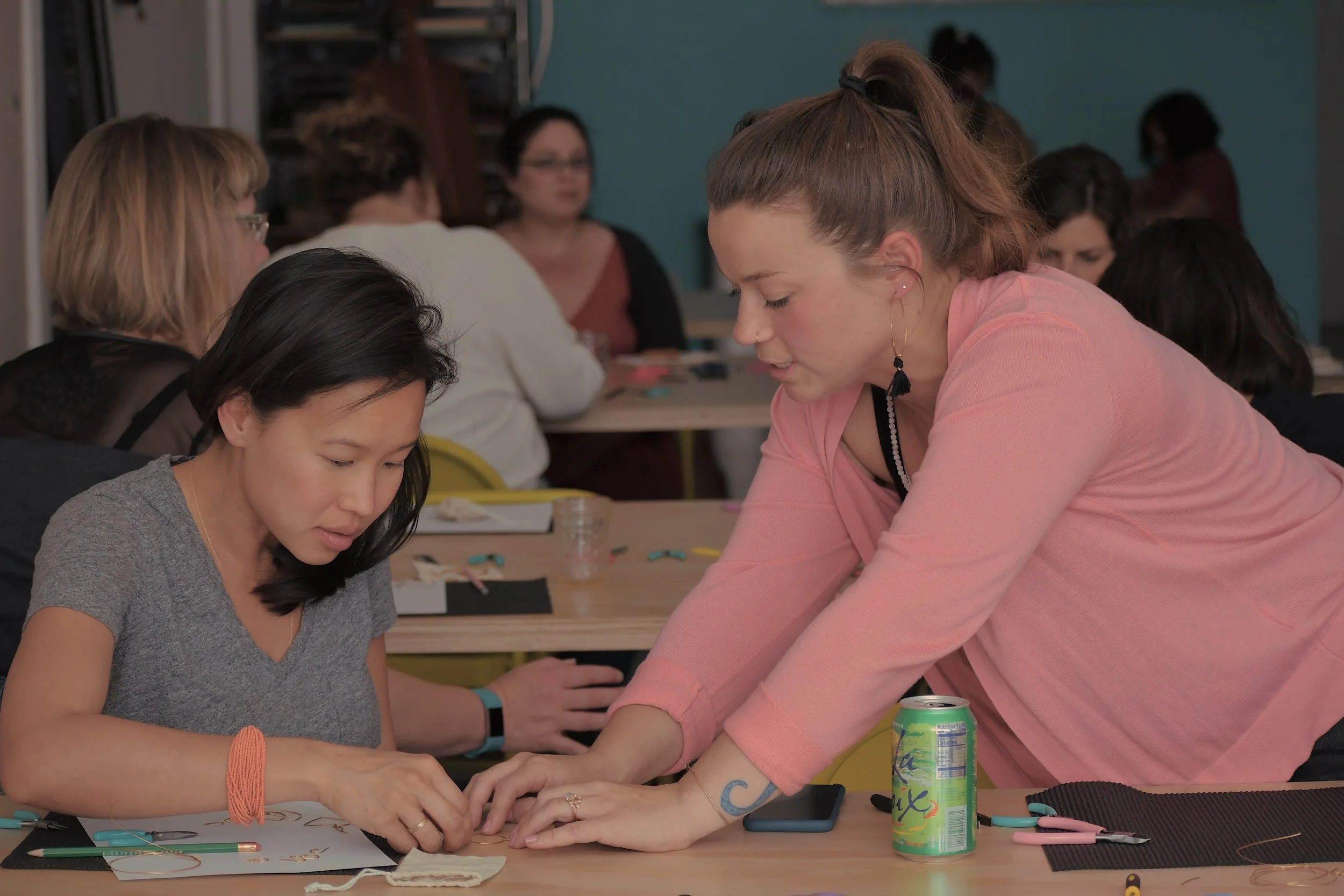 A group of women gathered around a table, engaging in a collaborative activity, with one woman standing and assisting another seated woman, in a casual indoor setting.