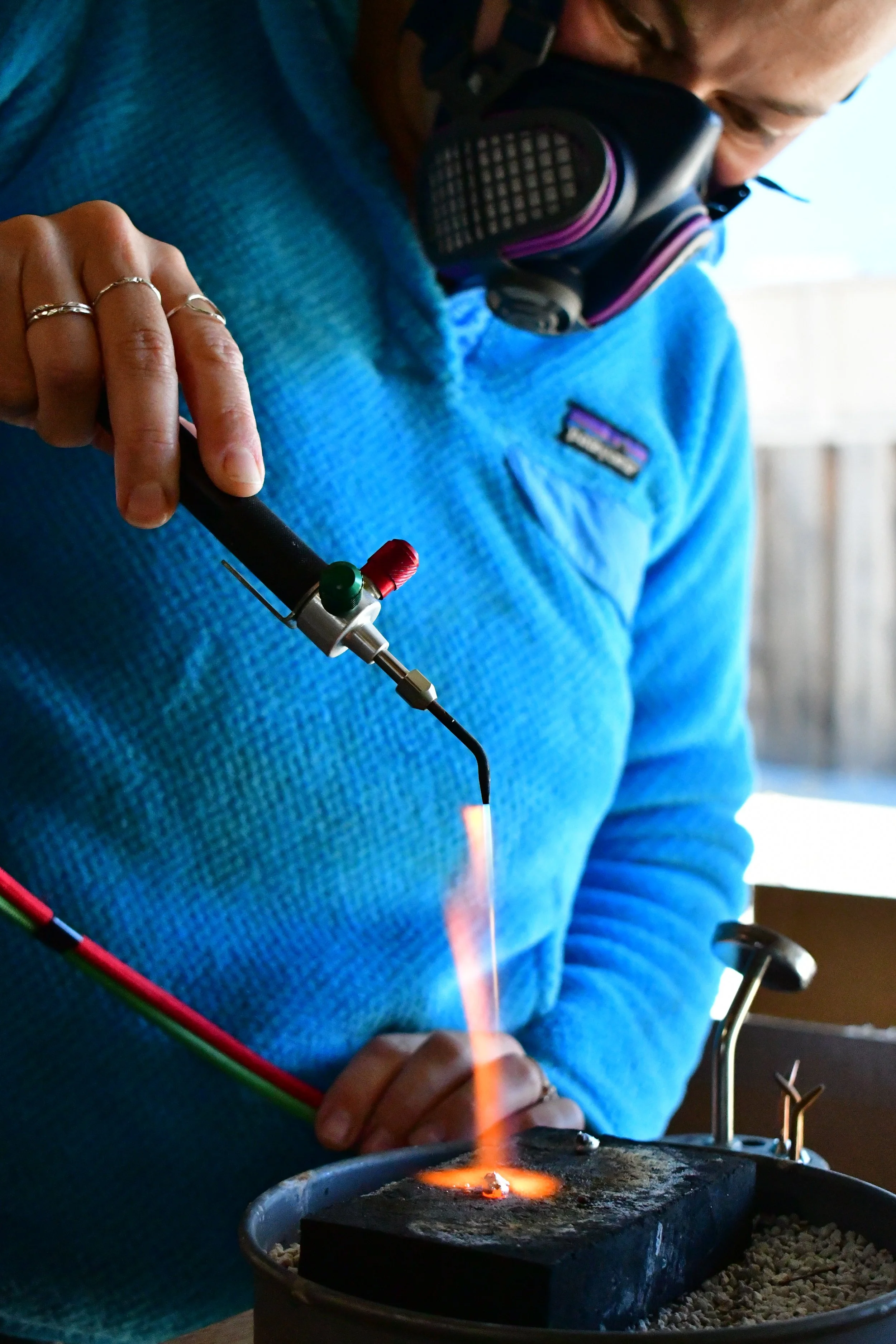 A person wearing a blue Patagonia fleece, with four rings on their fingers, is using a plasma cutter to cut metal. The plasma cutter produces a bright arc of light, and the person wears a respirator mask for safety. The workspace includes a container with gravel or small stones underneath the metal being cut.