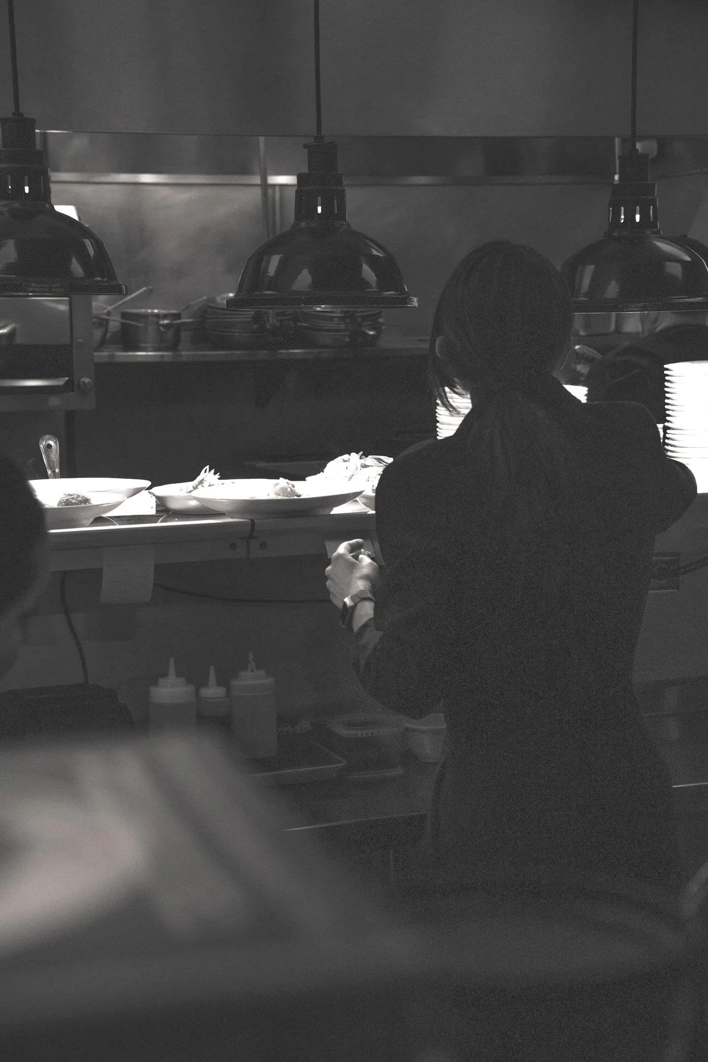 A person with long hair sitting at a counter in a restaurant or kitchen, with plates of food and condiment bottles in front of them, and hanging pendant lights overhead.