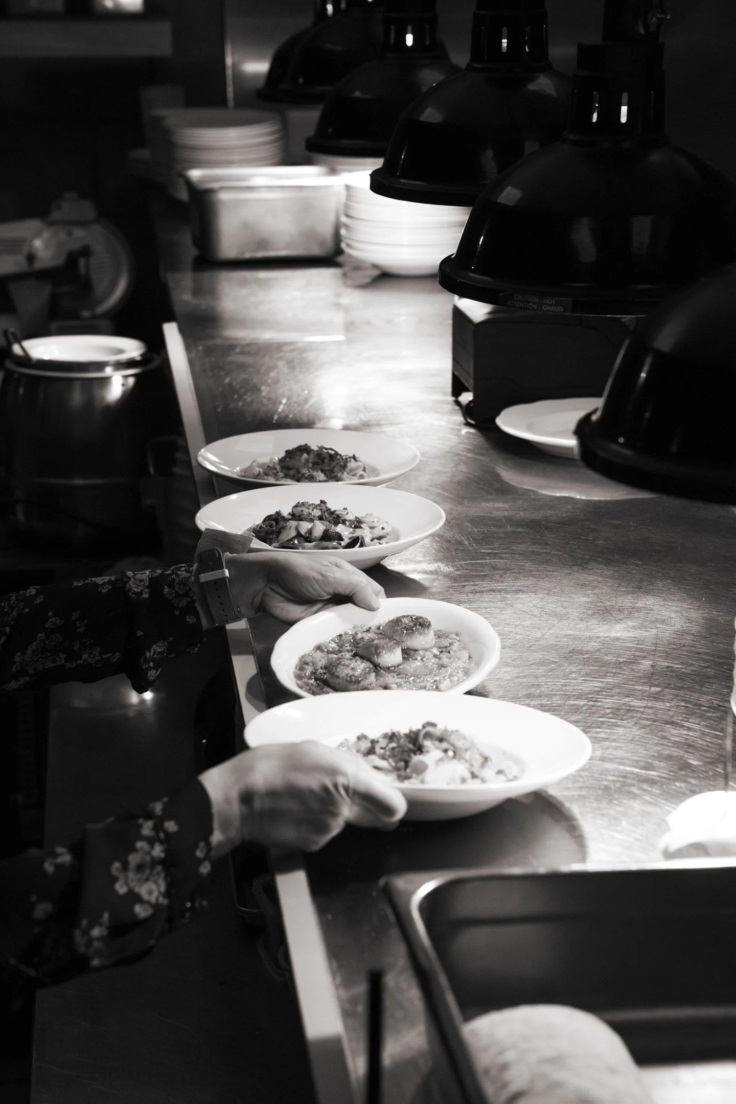 A restaurant kitchen window pass with trays of cooked food and bowls waiting to be served, illuminated under hanging heat lamps.