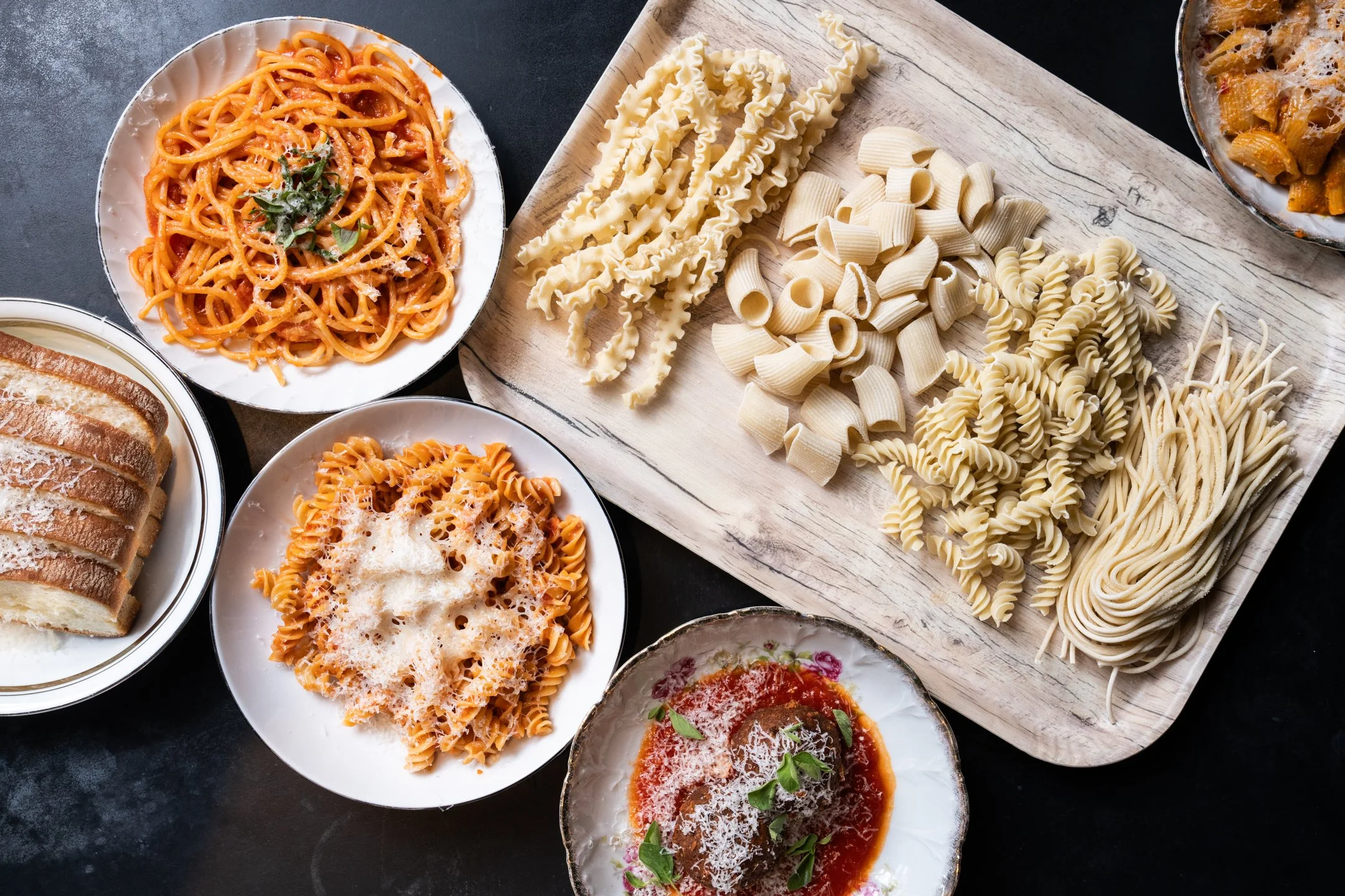 Assorted uncooked pasta varieties on a wooden cutting board, with plates of cooked pasta including spaghetti and fusilli, and a dish of baked pasta with cheese and tomato sauce.