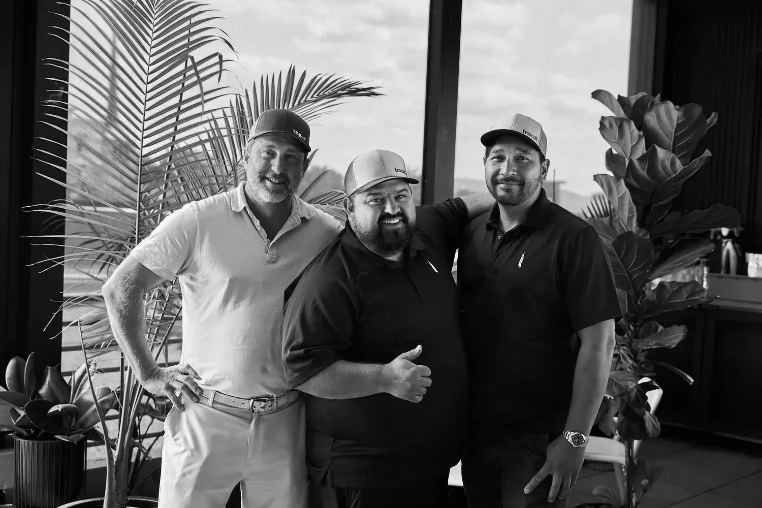 Three owners of Travail Collective wearing hats in a modern indoor space with large plants and a window in the background.