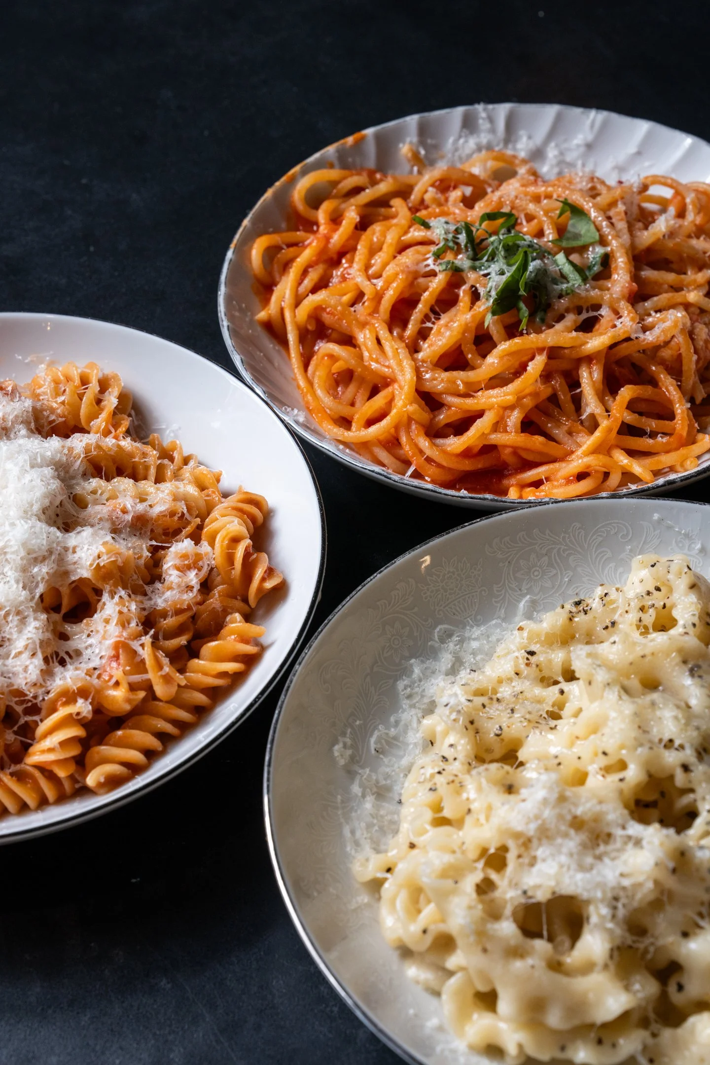 Three dishes of pasta on a black table, including spaghetti with tomato sauce, fusilli with cheese, and macaroni with cheese and pepper.