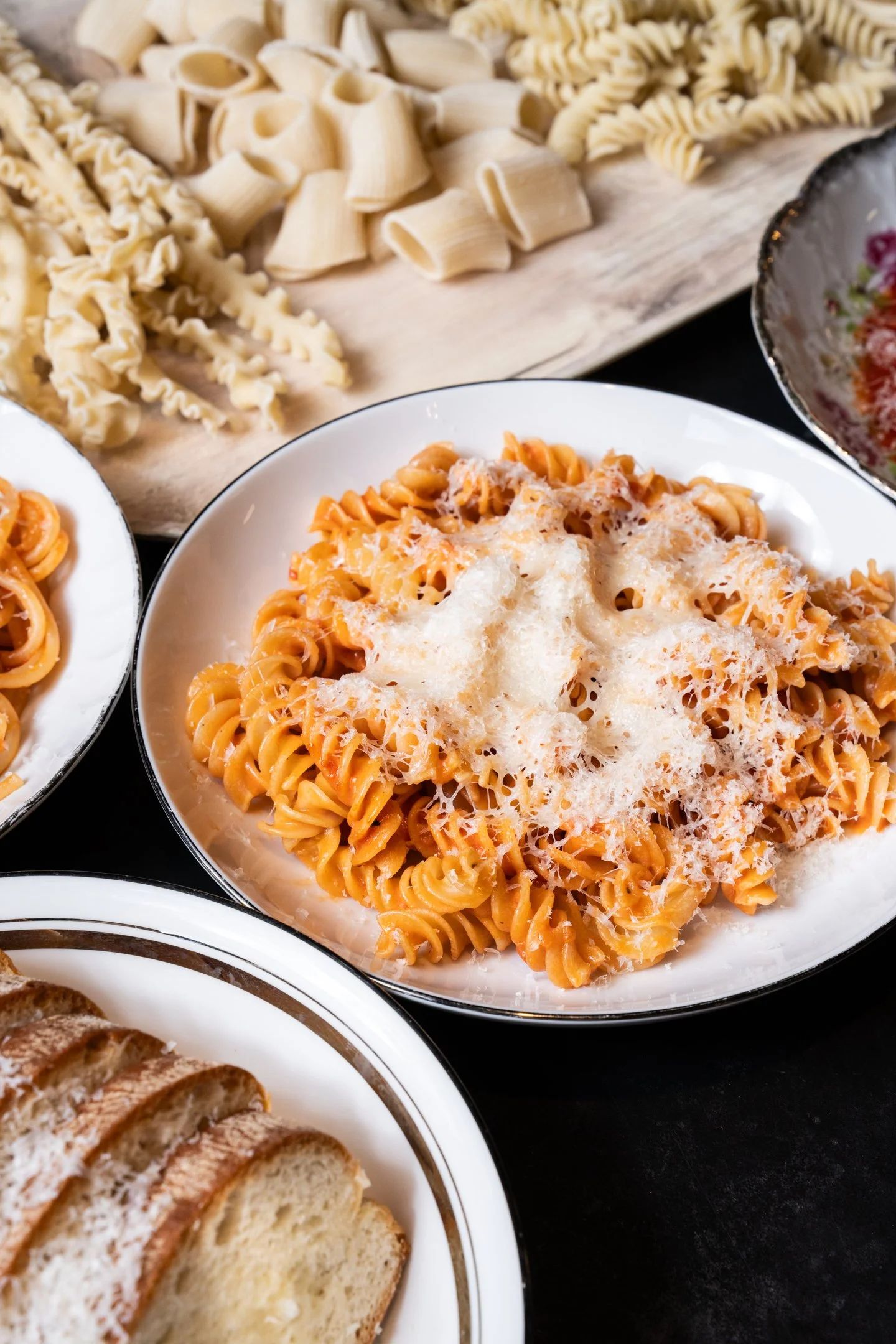 A plate of pasta with tomato sauce and grated cheese, and a wooden board of uncooked pasta in various shapes.