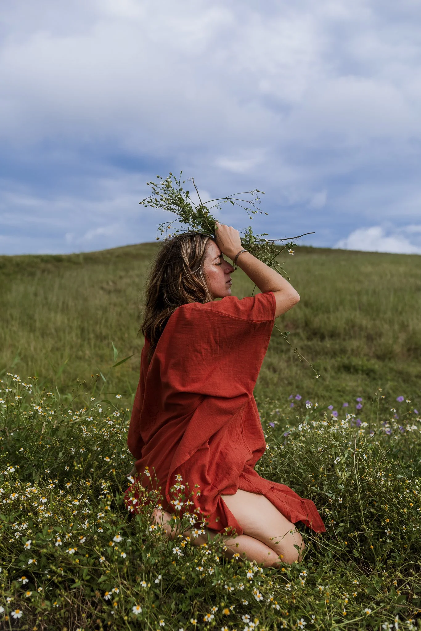 A woman in a red dress sitting in a grassy field filled with small white and yellow flowers, holding a bouquet of wildflowers on her head, with a cloudy sky in the background.