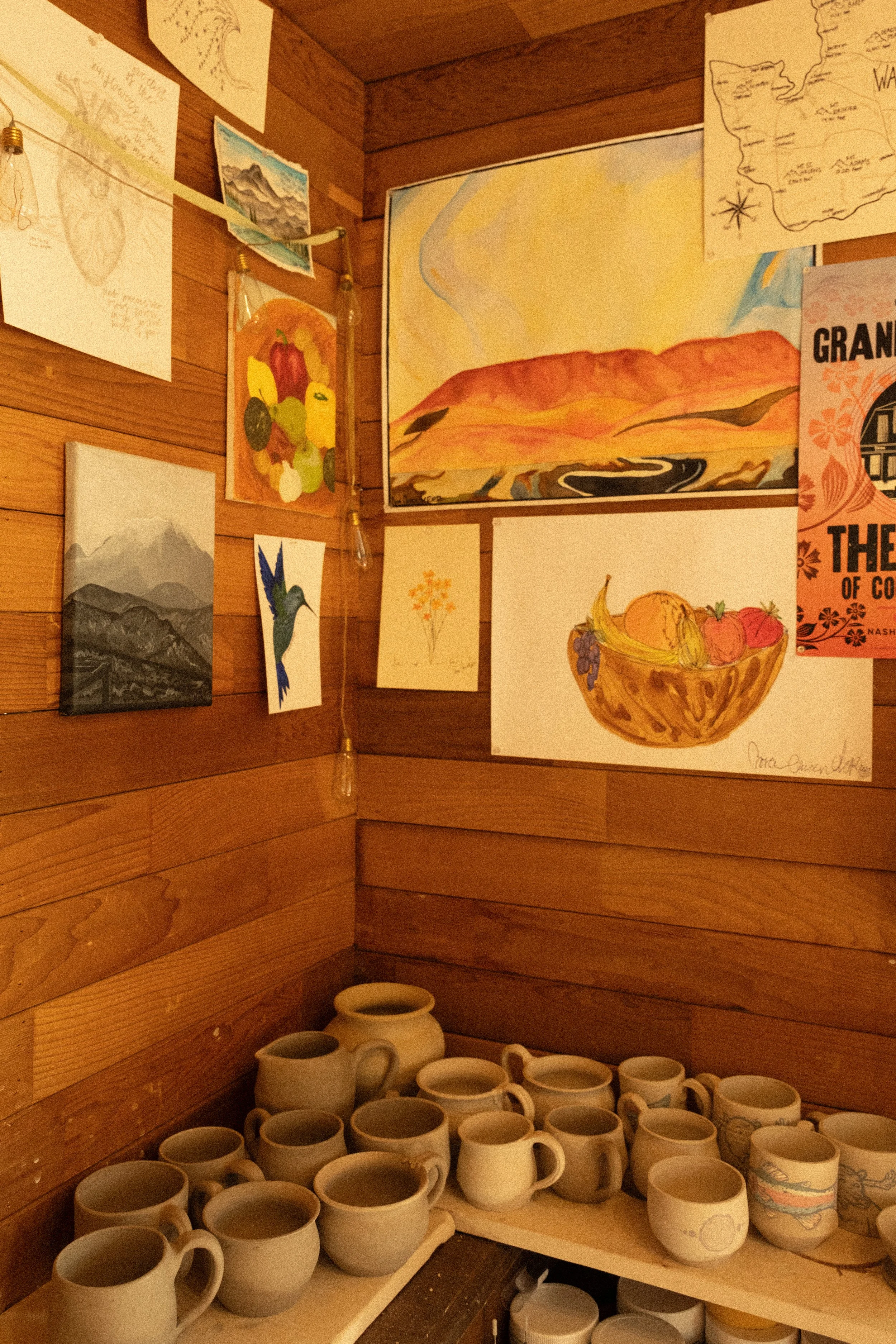 Display of unfinished ceramic mugs and pottery at a craft studio with artwork and maps on wooden walls.