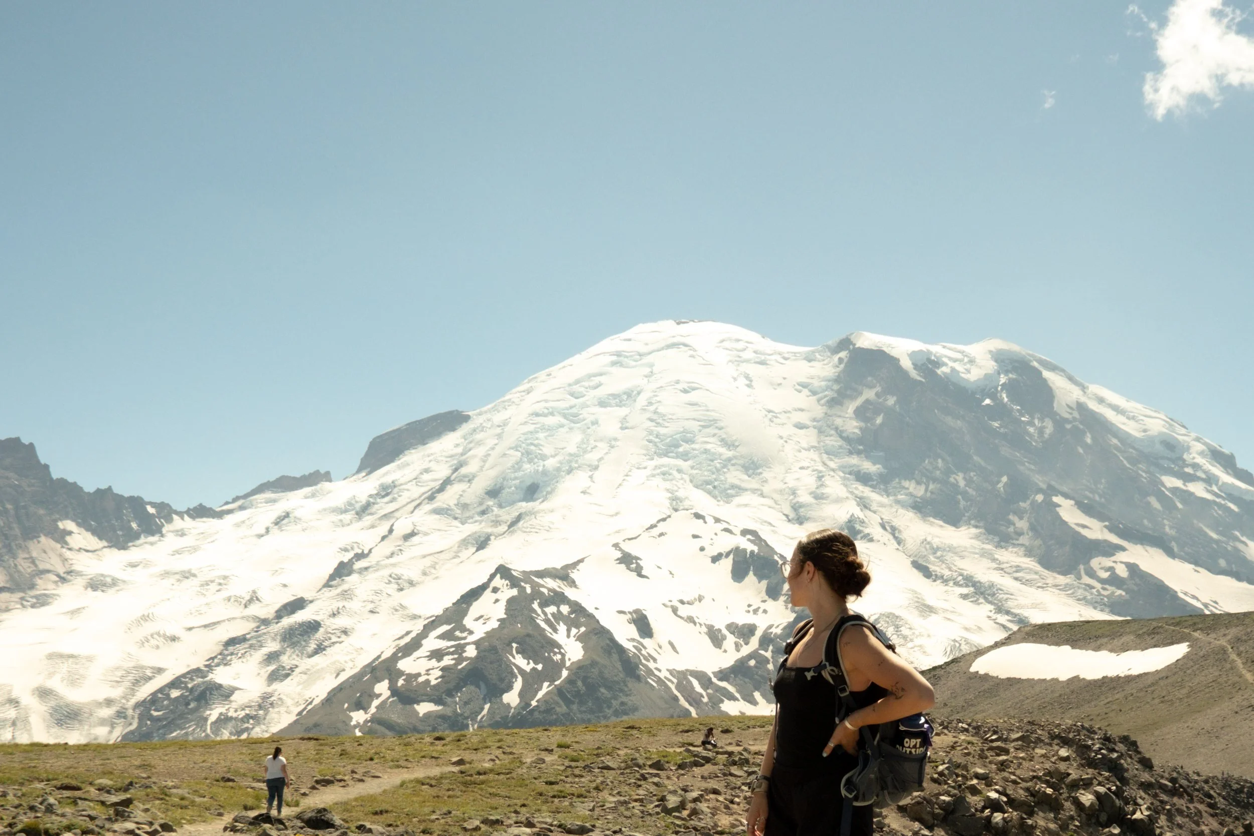 A woman with sunglasses and a backpack standing in front of a snowy mountain landscape.