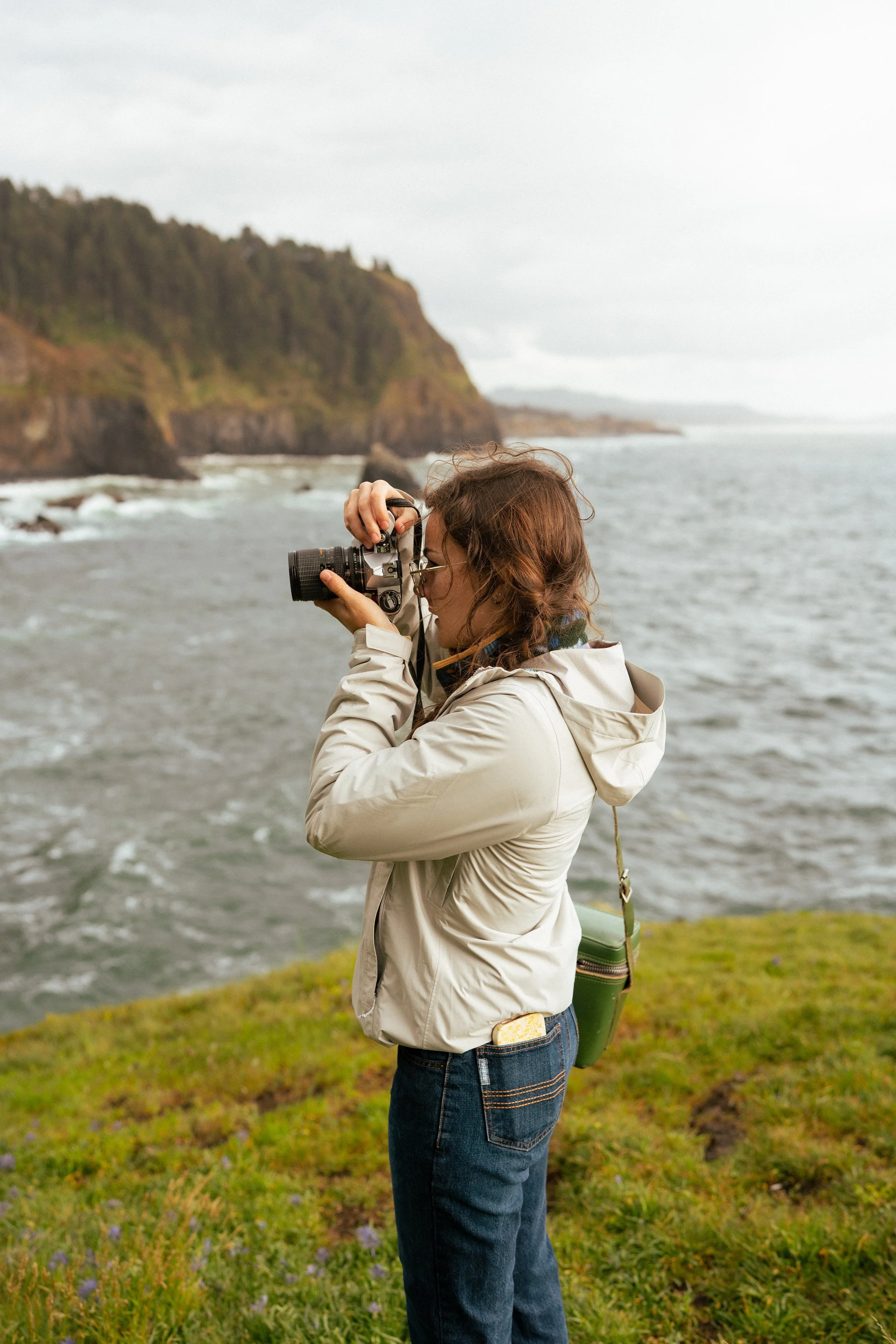 Woman in beige jacket and jeans taking a photo with a camera near the coast, green hills and cliffs in the background.