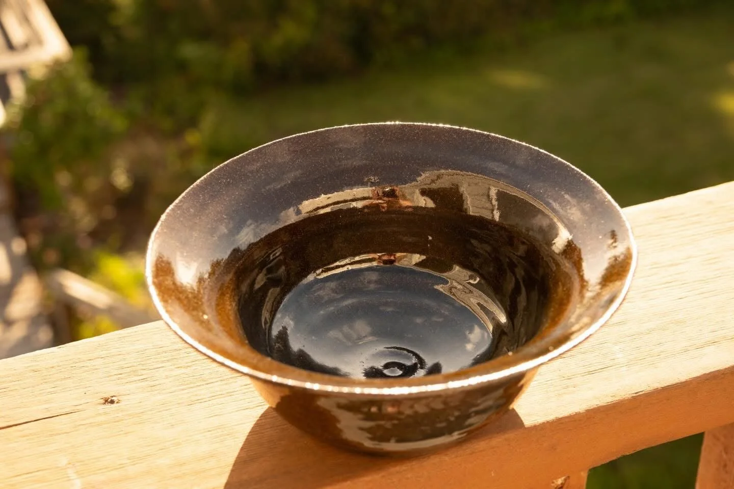A small, round ceramic bowl with a dark glaze, placed on a wooden surface outdoors, reflecting the surrounding sky and trees.
