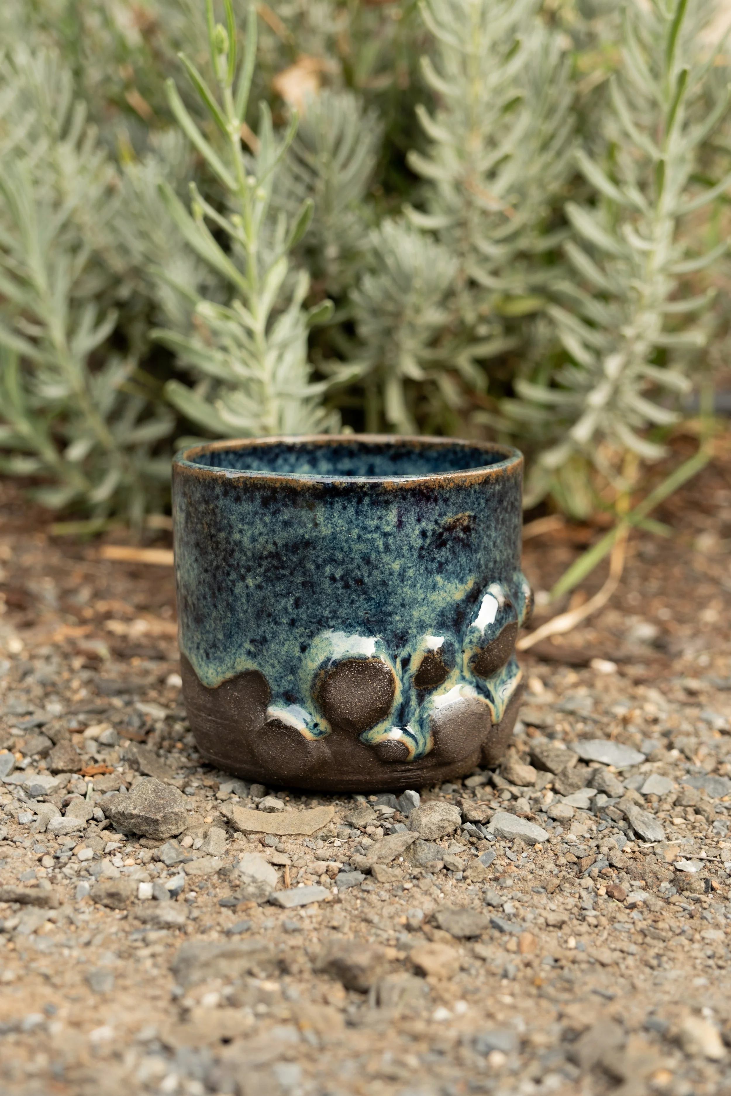 A ceramic mug with a black, blue, and white glazed top sits on gravel, with green plants in the background.