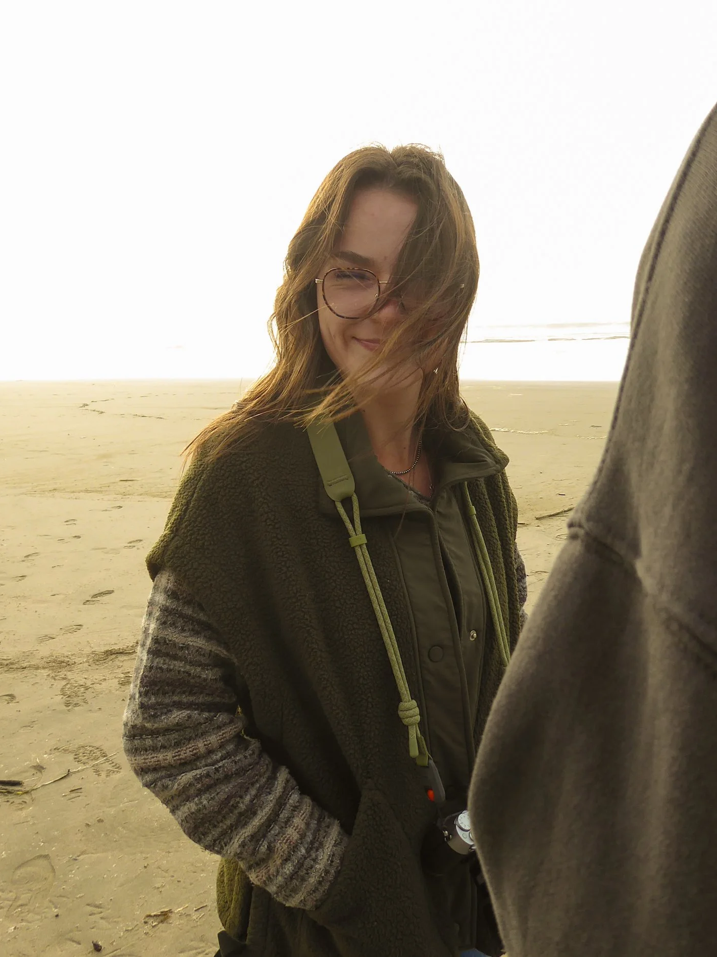 A woman with curly light brown hair, wearing glasses, a green jacket, and a brown fleece coat, smiling at a beach.