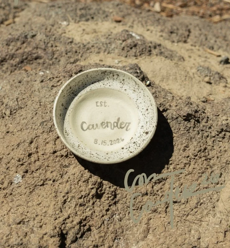 Small round ceramic ring dish with handwritten words "Est. Cavender 8.15.2026" inside, placed on dirt ground outdoors with sunlight.