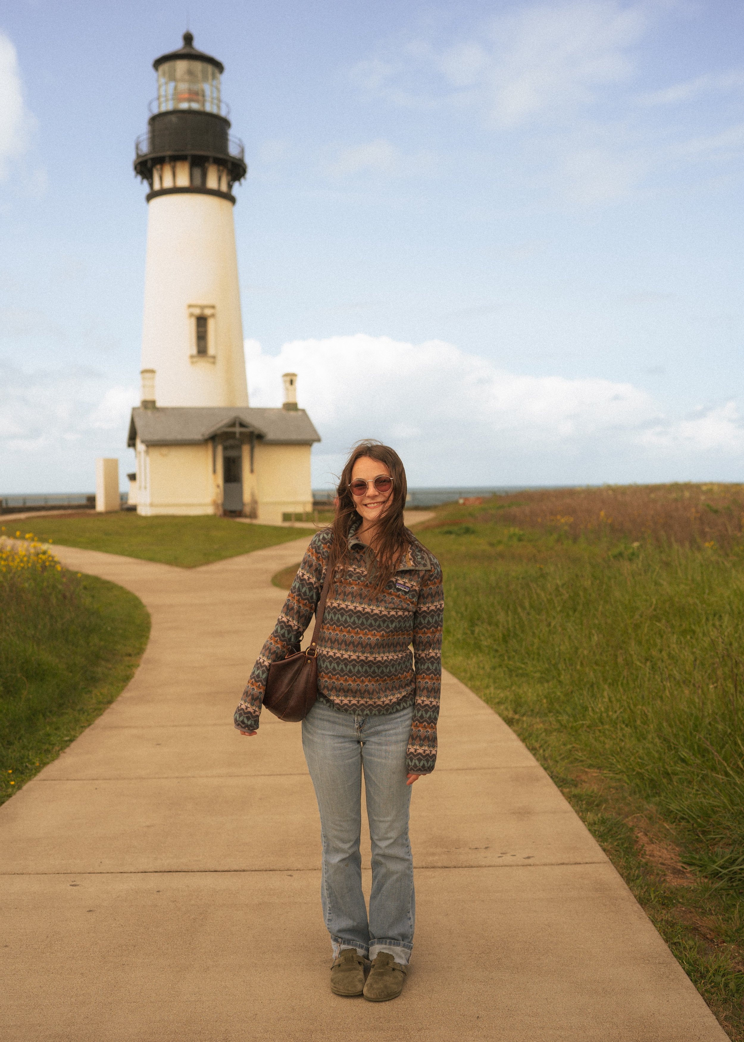 A woman with long hair wearing sunglasses, a patterned jacket, light blue jeans, and brown shoes, smiling and standing on a paved path near a lighthouse with a blue sky and some clouds in the background.