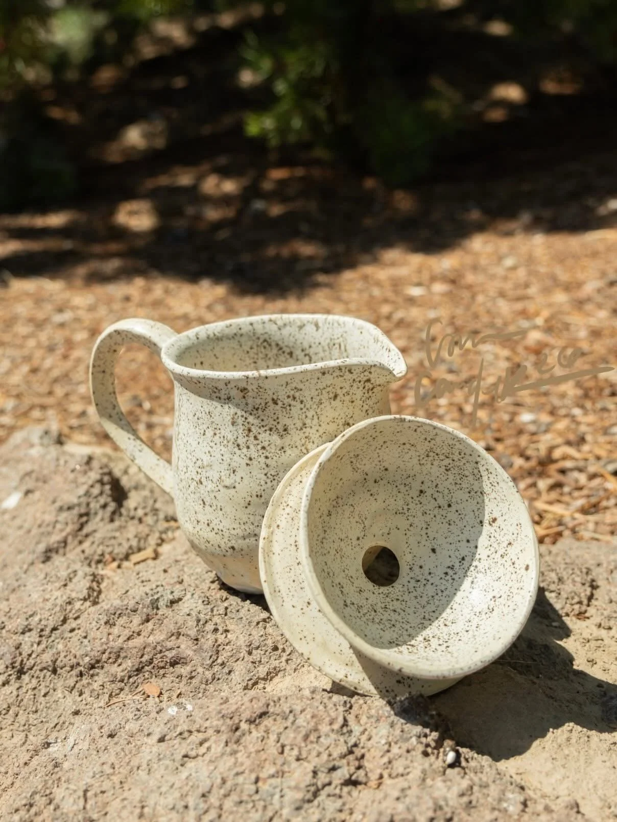 A ceramic coffee pour over with a speckled white and brown glaze, placed on a rocky surface outdoors with a blurred background of trees and soil.