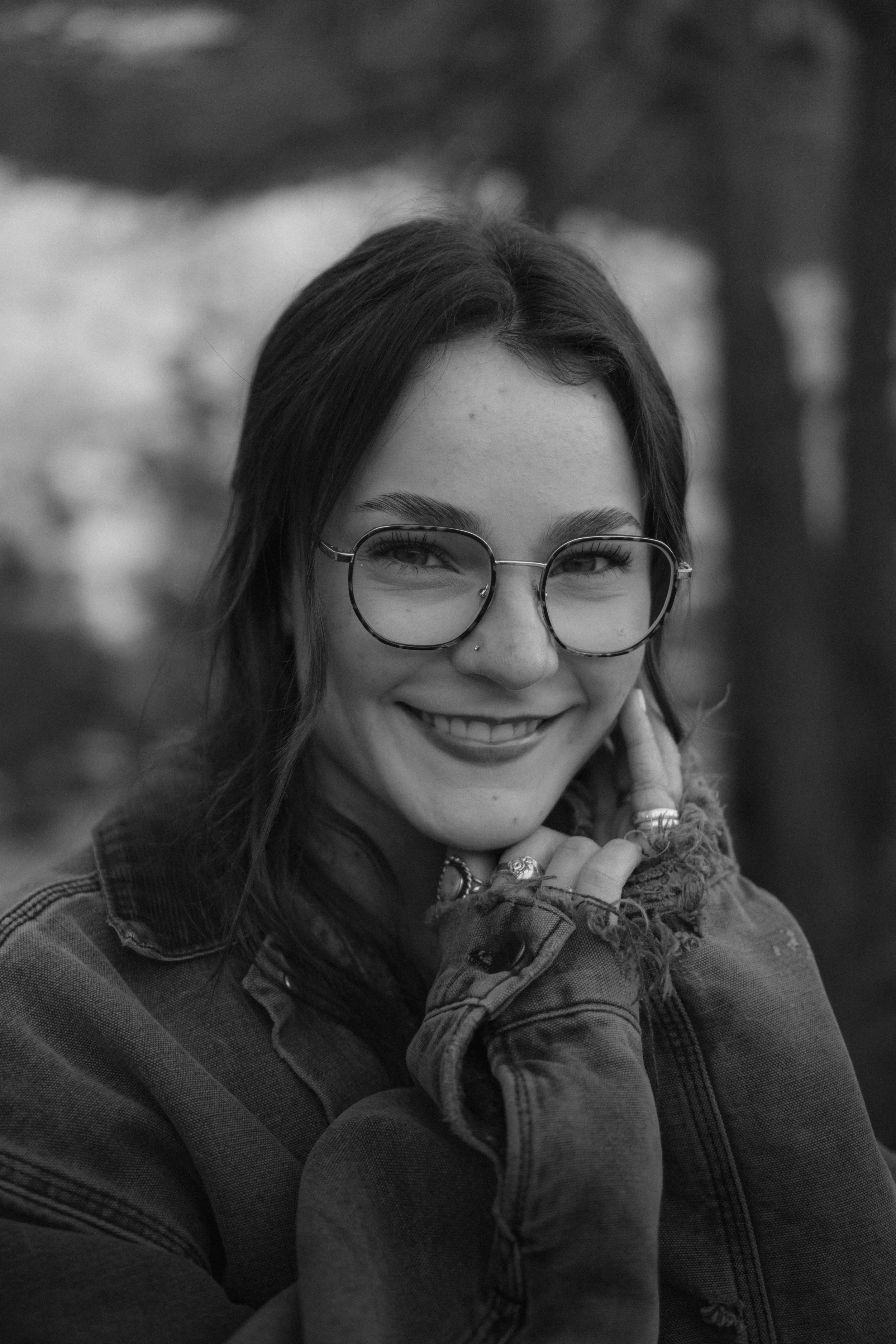 Black and white photo of a young woman with long hair, glasses, and a nose piercing, smiling and resting her chin on her hand, outdoors in a park or wooded area.