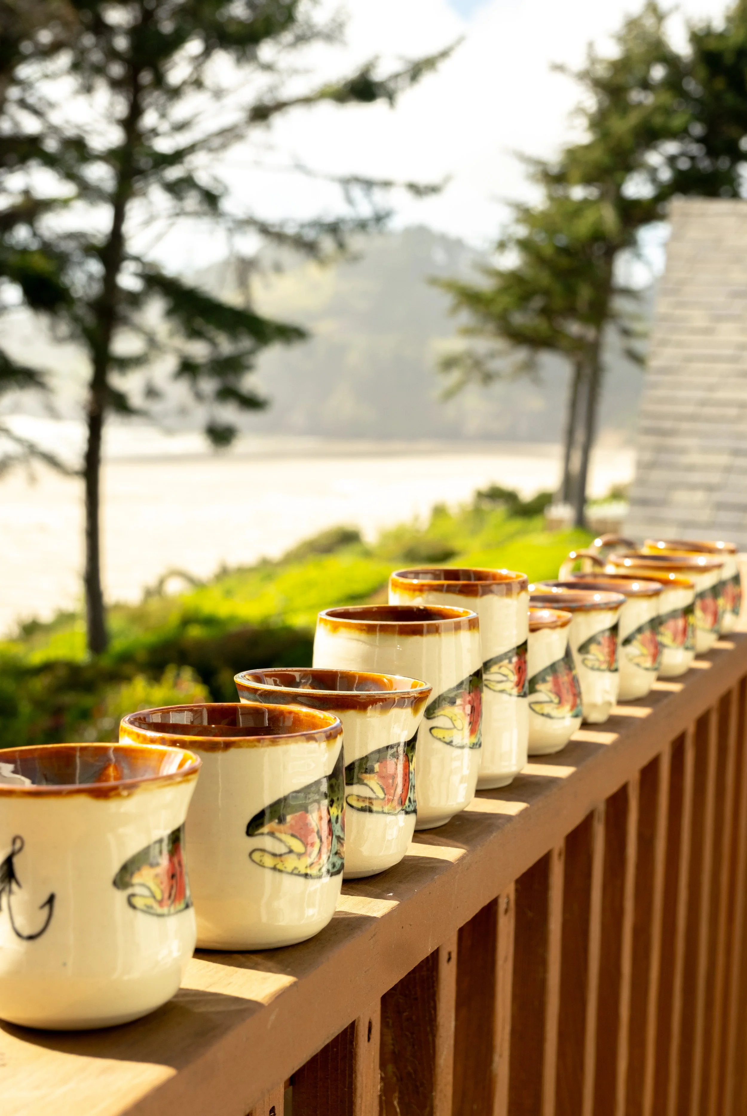 Row of ceramic mugs with colorful fish designs on a wooden railing outdoors, with trees and a building in the background.
