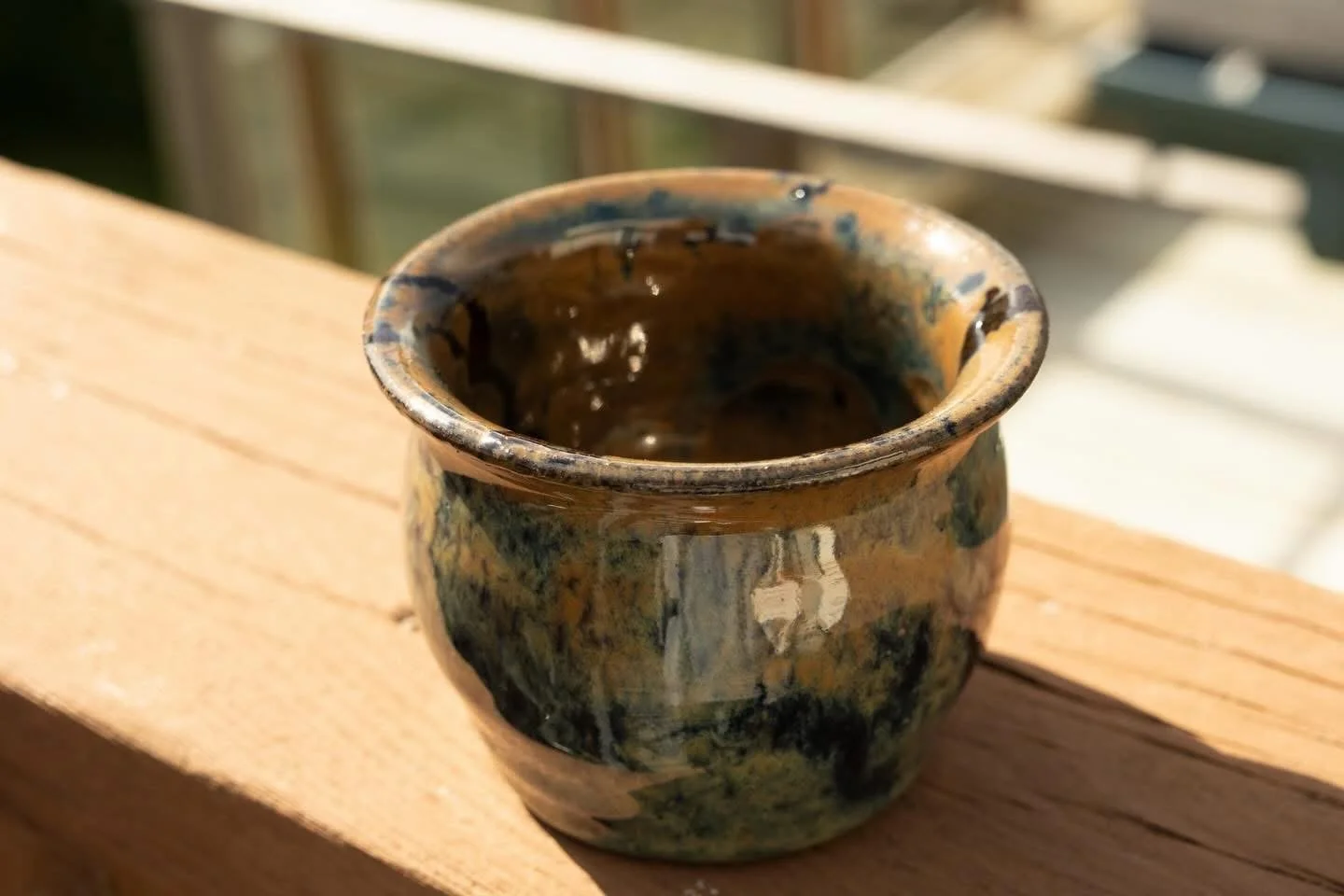 A small ceramic pot with a glossy, multicolored glaze, placed on a wooden surface outdoors.