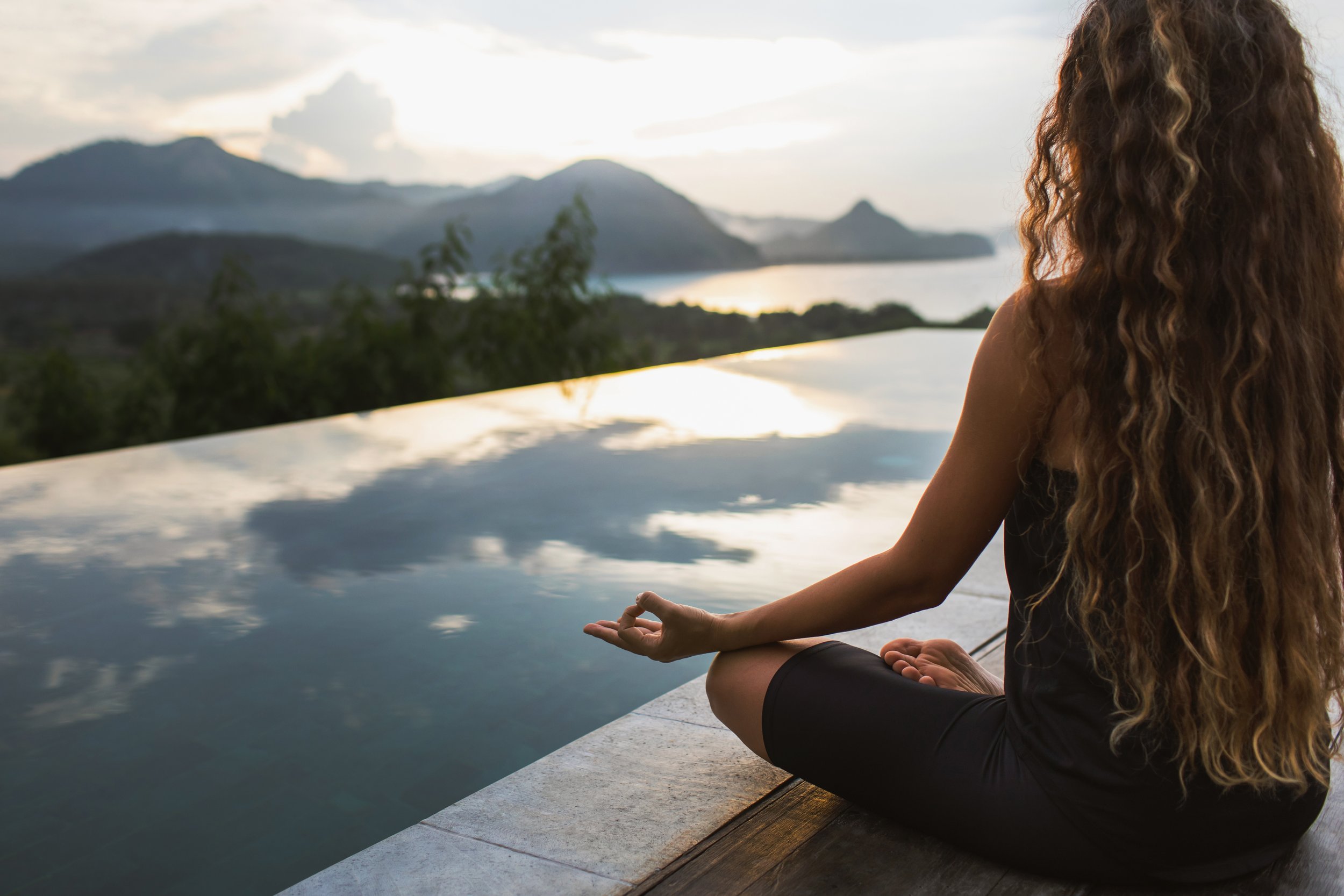 A woman with long curly hair sitting cross-legged on an outdoor surface near a pool, meditating with eyes closed, overlooking a scenic mountain and lake landscape at sunrise or sunset.