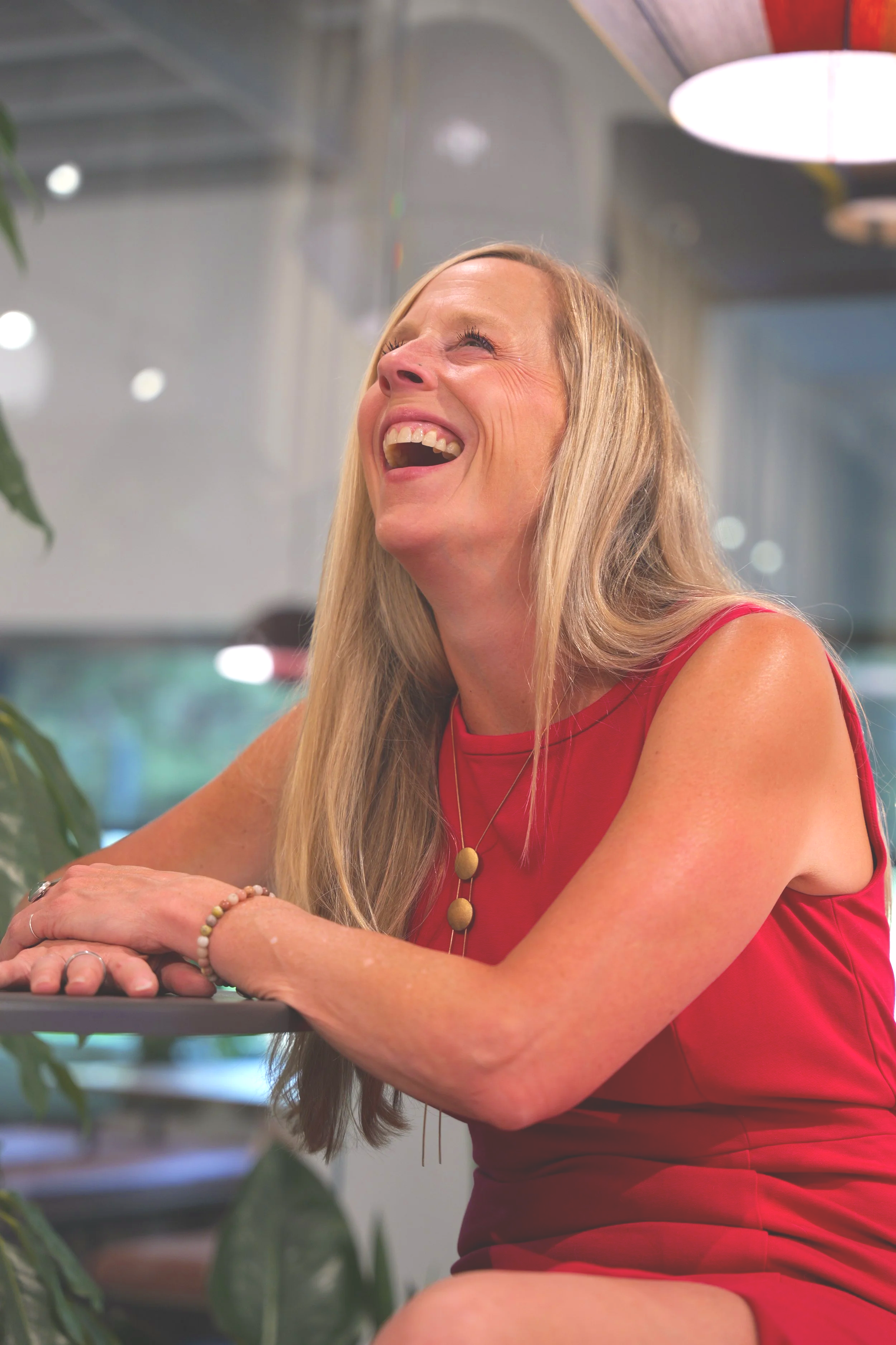 A woman with long blonde hair laughing and smiling while seated at a table in a room with modern decor, wearing a sleeveless red top and jewelry, with blurred background and greenery.