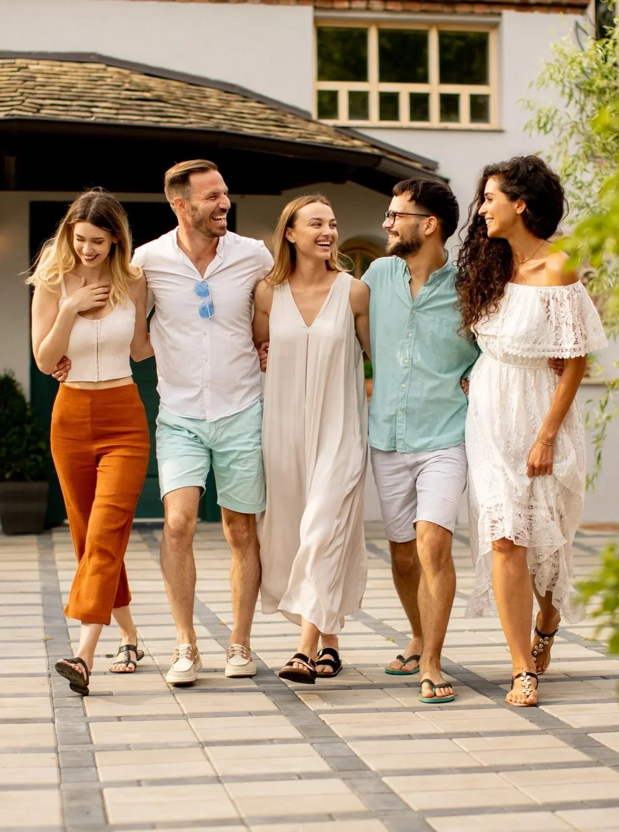 A group of five friends walking arm-in-arm outside on a sunny day, smiling and laughing together.