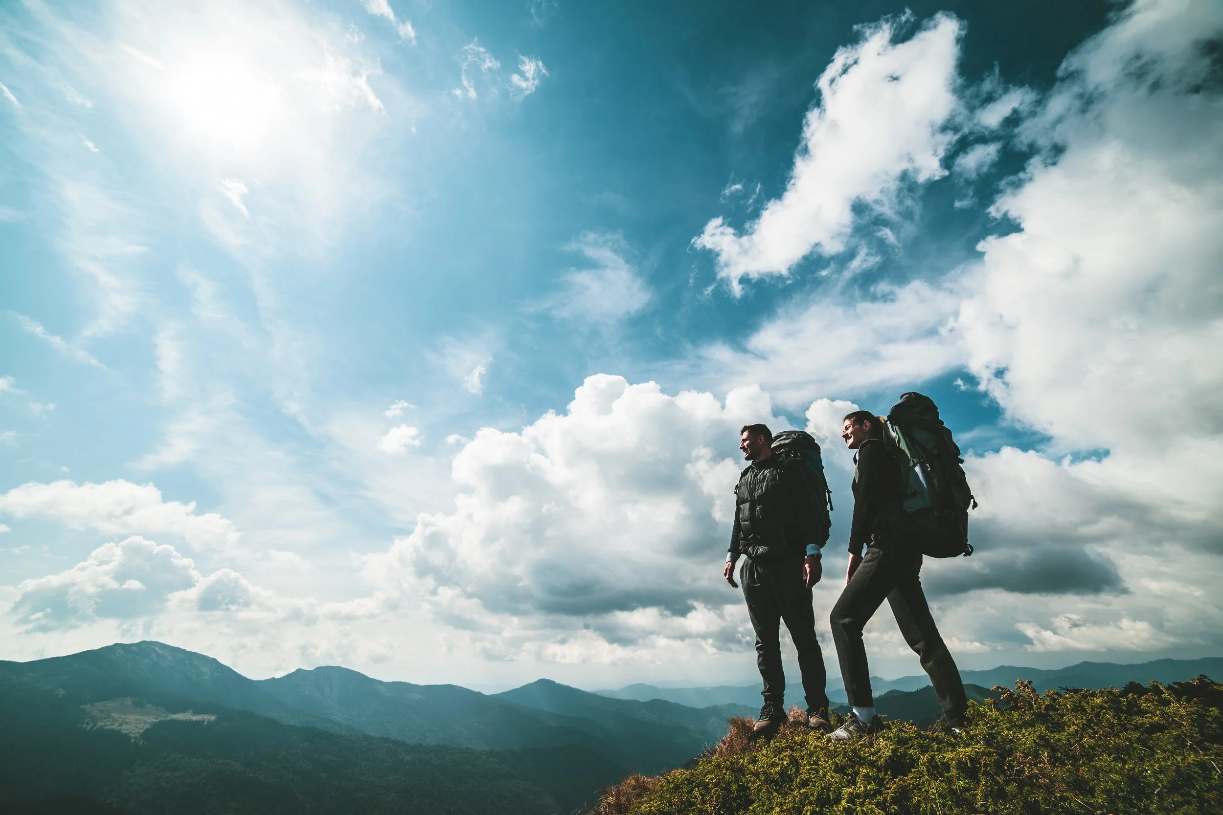 Two hikers with backpacks walking on a mountain trail under a partly cloudy sky, with distant mountain ranges in the background.