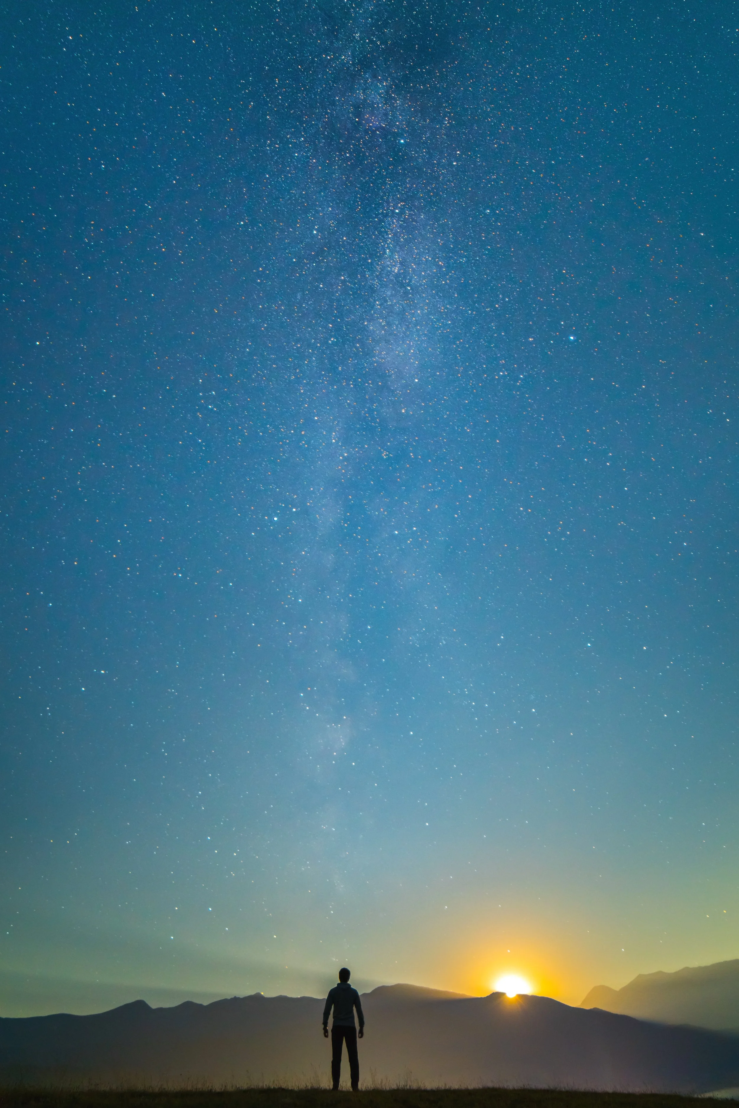 A person stands in a field, gazing at a starry night sky with the Milky Way visible, mountains in the distance, and a sunset or sunrise on the horizon.