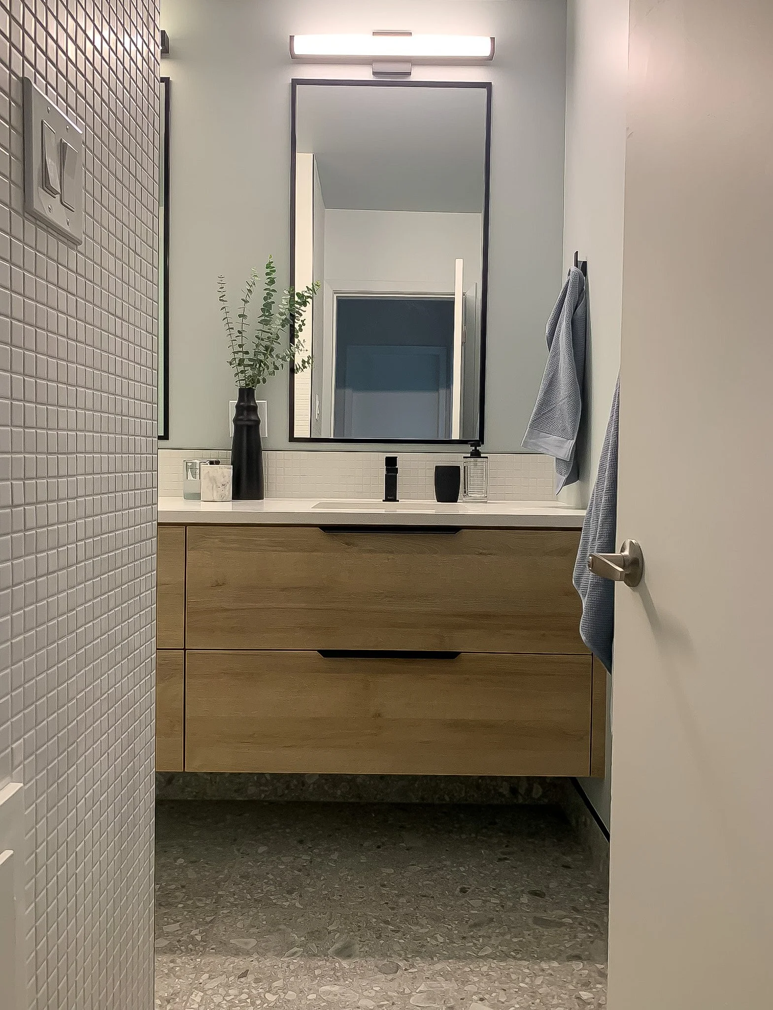 A modern bathroom with a large mirror above a wooden vanity with a black faucet, decorated with a black vase holding green foliage and a small vase. To the right, a towel hangs on a hook. The bathtub or shower wall has a white tile pattern, and the f