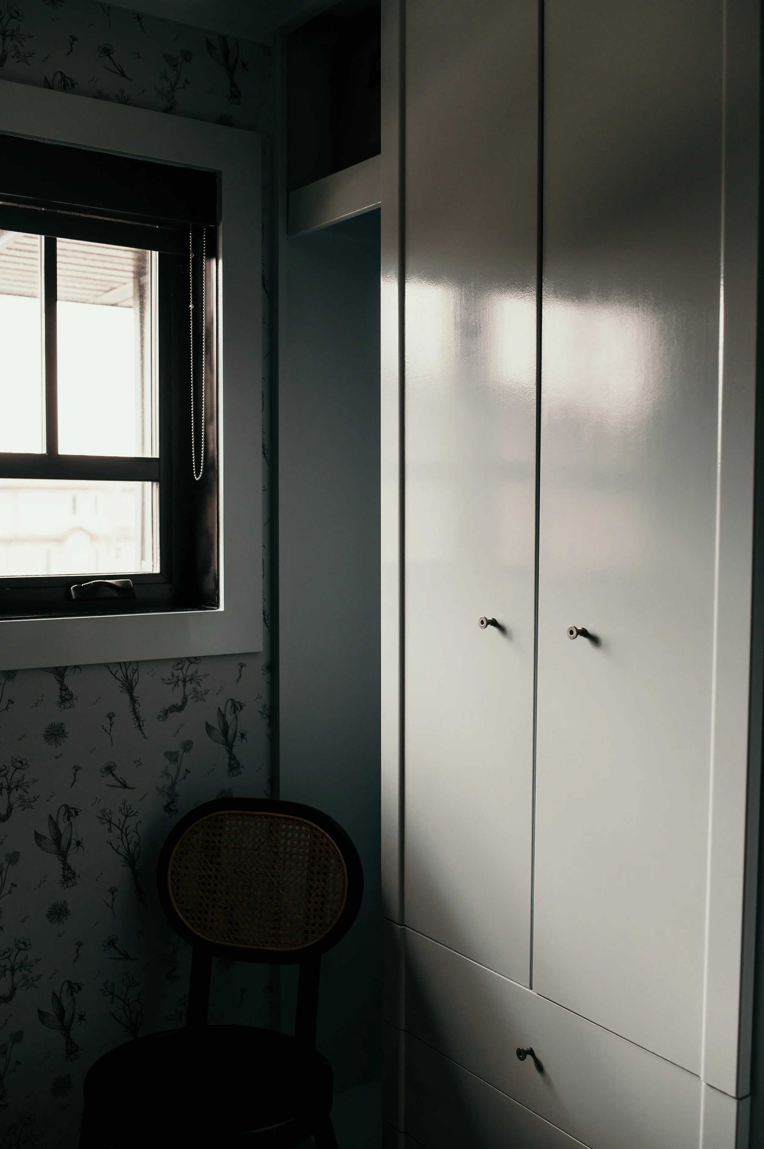 A white wardrobe with black handles standing next to a window with dark blinds in a room with floral wallpaper. There is a woven chair with a round seat and black legs below the window.