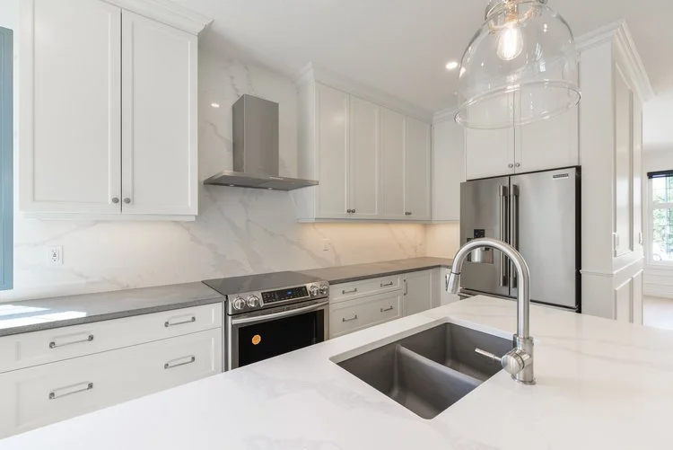 Modern white kitchen with marble backsplash, stainless steel refrigerator, oven, and a large white island with a sink and a curved faucet.