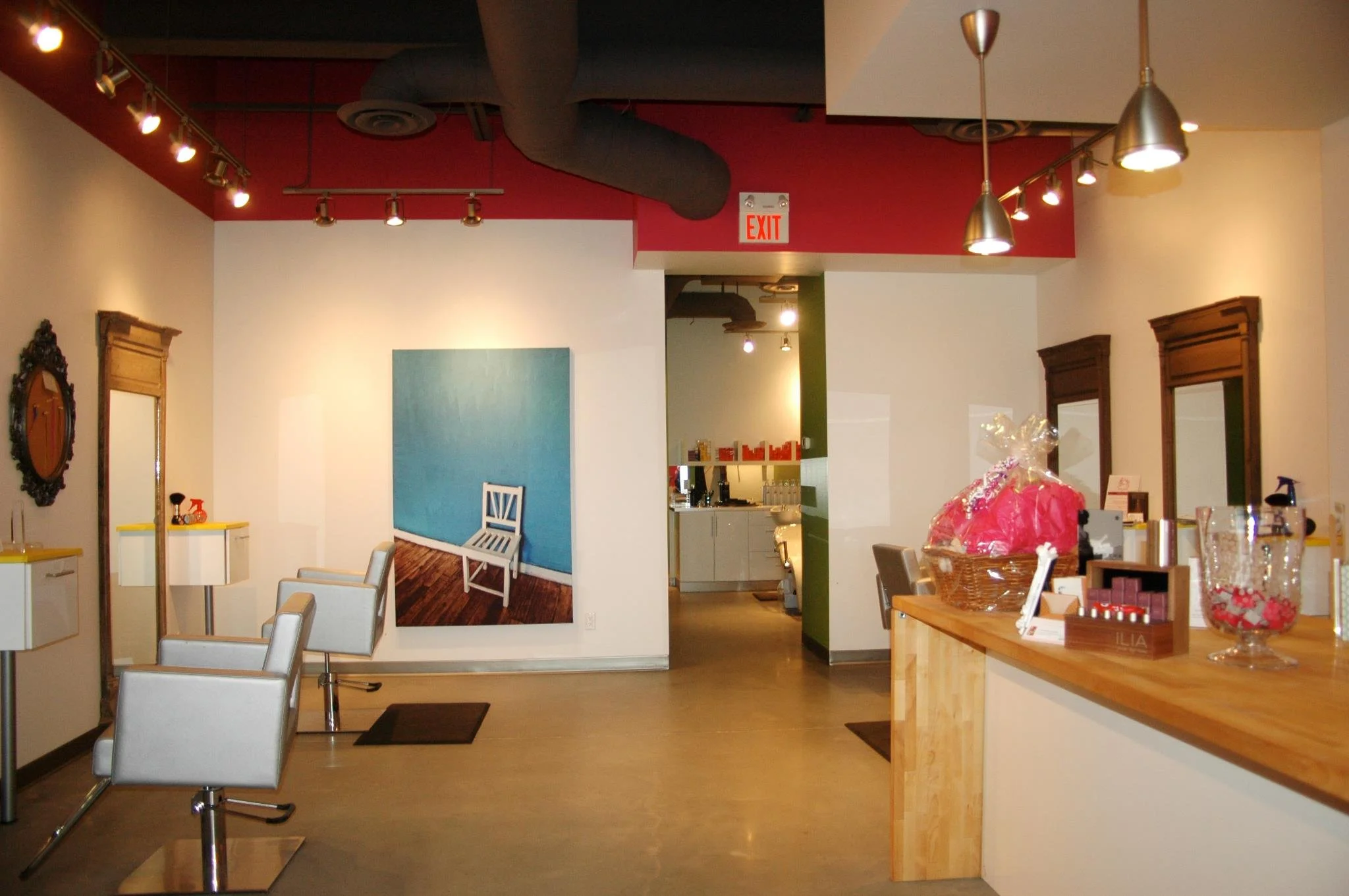Interior of a modern cafe or restaurant with white dining chairs, wooden tables, decorative mirrors, colorful paintings, and a dessert display with baked goods wrapped in clear plastic on a counter.