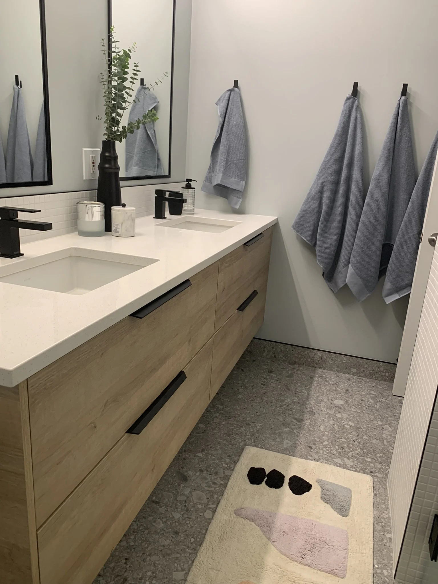A modern bathroom with a double vanity with light wood cabinets and white countertops featuring two sinks, black faucets, and decorative items including a tall black vase with green foliage, pump soap dispenser, and candles. There is a large mirror a
