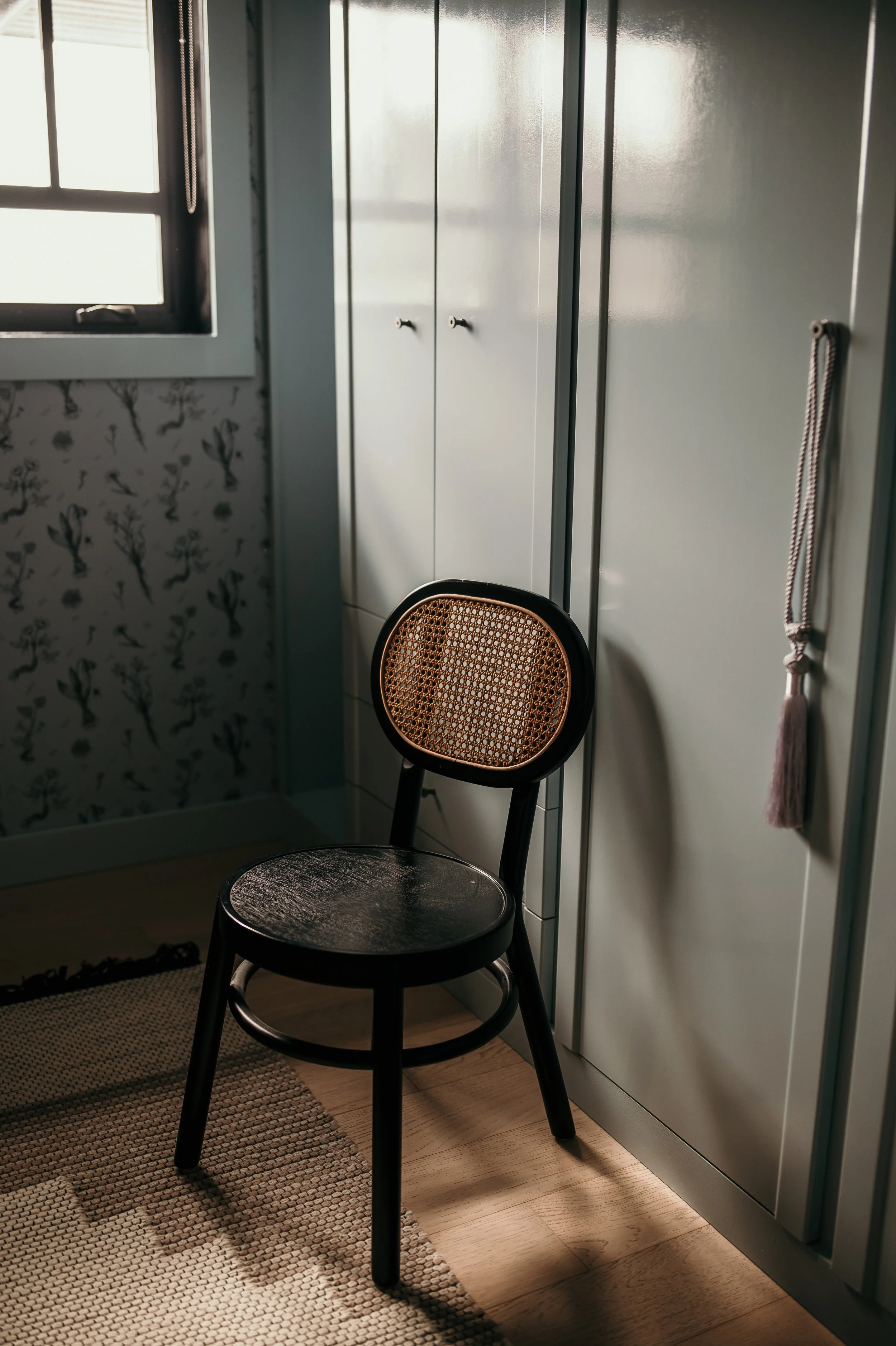 A black chair with a woven rattan backrest and round seat in a room with a patterned wall and gray wardrobe with handles, next to a window with a cord hanging on the side.