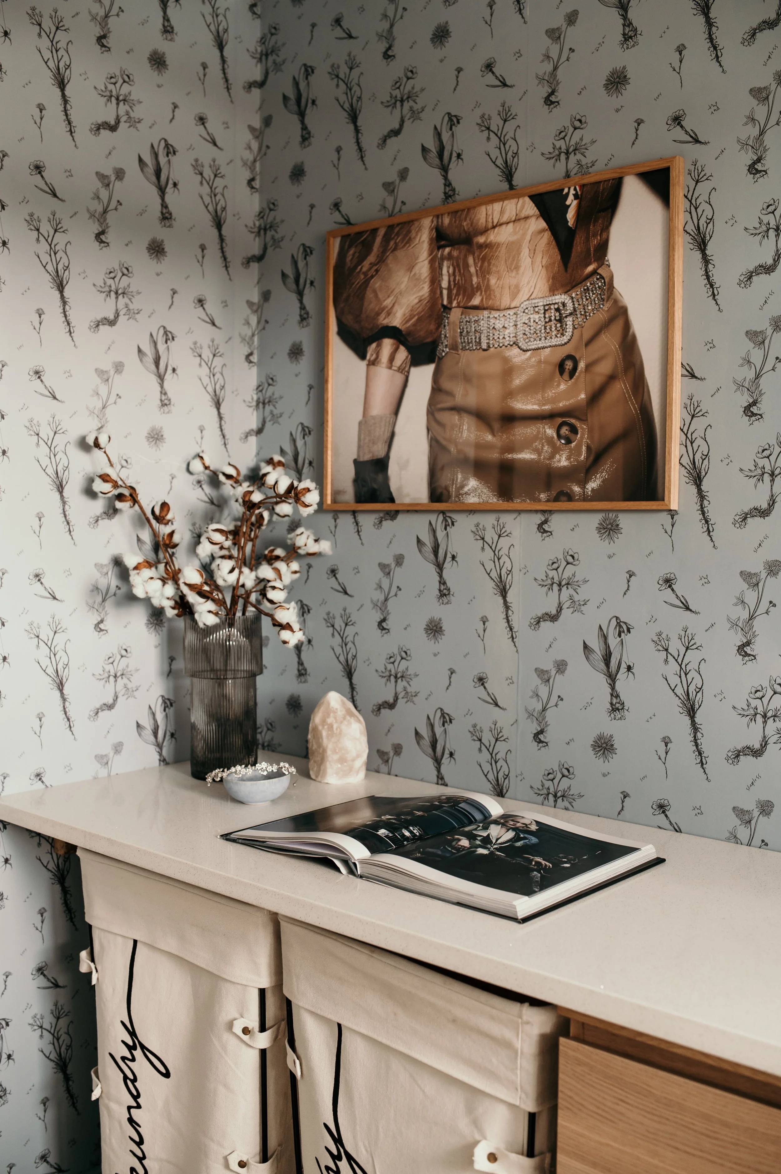 A corner of a room with a decorative wallpaper featuring black floral sketches on a light background. There is a framed photograph of a woman wearing a tan outfit and sparkling belt hanging on the wall. A white table below the photo holds a vase with