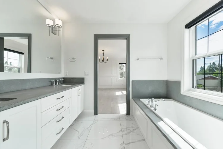 Modern bathroom with white walls, a large bathtub by the window, a gray countertop with a sink, and a mirror, with a view into an adjoining room with a chandelier and window.
