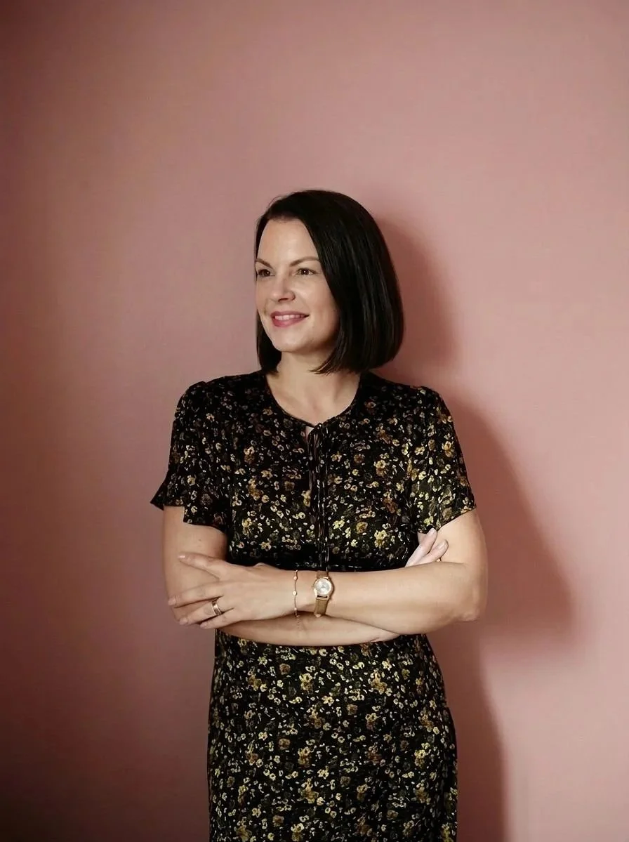 A woman with short black hair, wearing a black dress with gold floral patterns, standing with her arms crossed against a pink wall, smiling and looking to her left.
