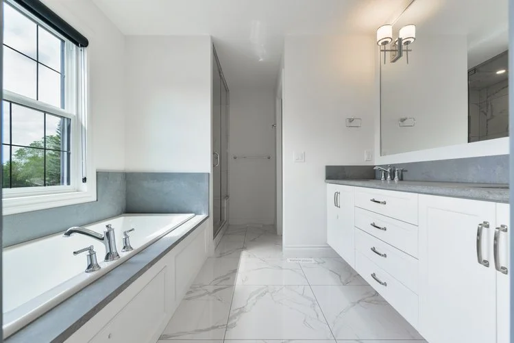 Modern bathroom with white cabinets, gray countertops, a bathtub under a large window, and a wall-mounted light fixture.