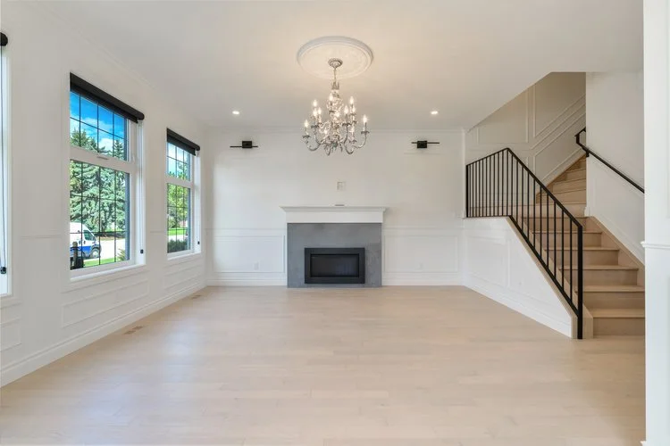 Empty living room with large windows, a modern fireplace, a chandelier, and a staircase with a black railing.