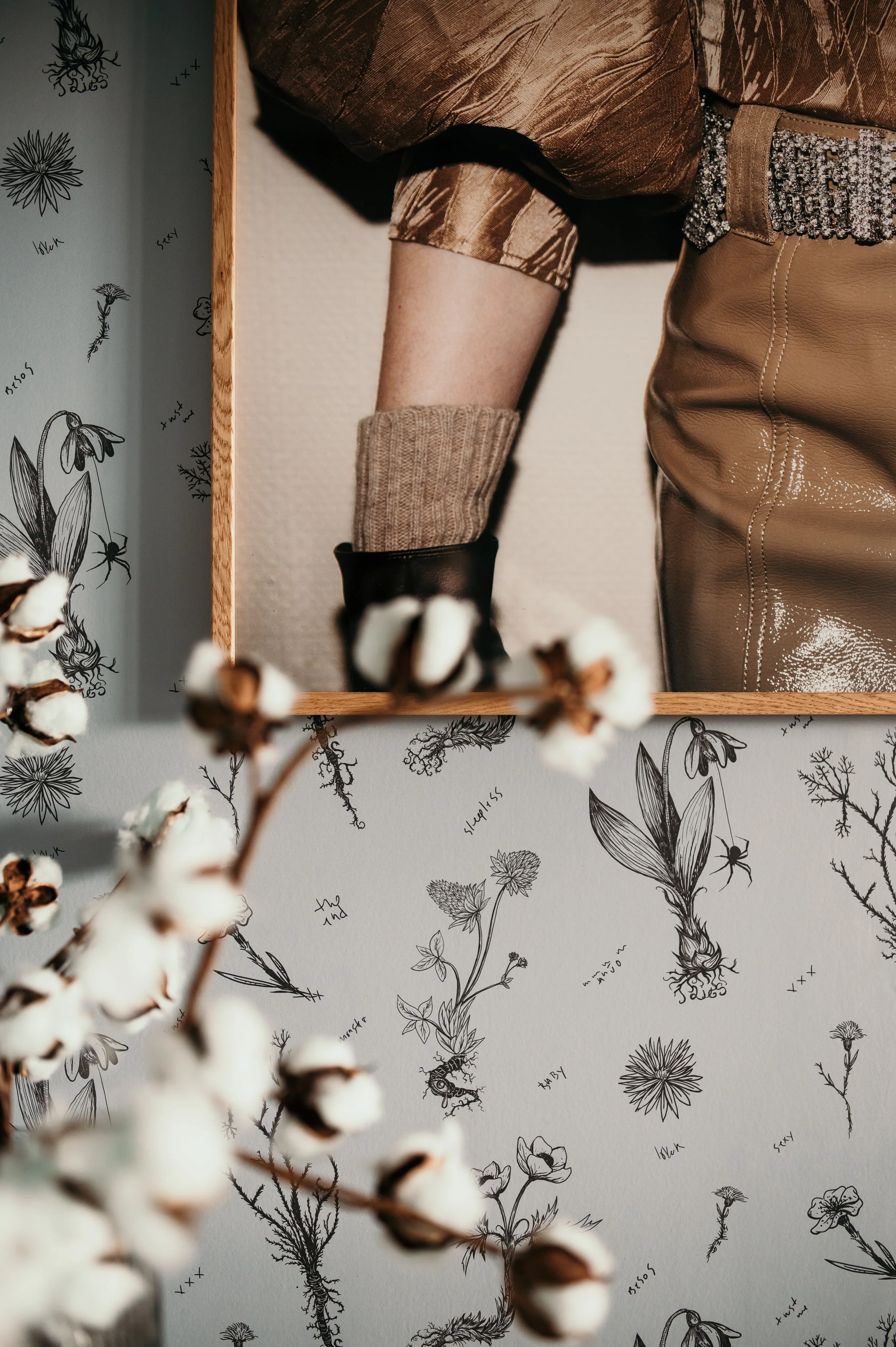 Close-up of a person wearing a metallic bronze jacket, beige knitted glove, and brown leather pants, standing in front of a wall with black botanical drawings and handwritten notes, with cotton branches in the foreground.