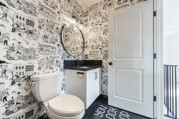 A small bathroom with black and white cityscape wallpaper, a white toilet, a white vanity with a black countertop and mirror, a black and white rug on the floor that says 'SOUR.'