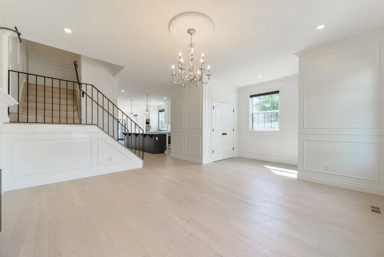 Bright, spacious living room with white walls, light wood flooring, a chandelier, and large windows.