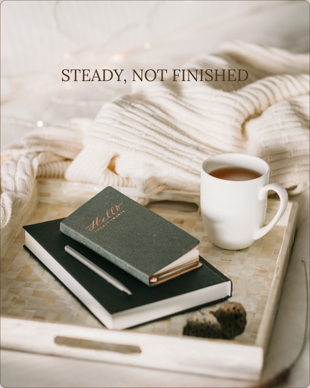 A tray with a cup of tea, two notebooks with a pen on top, and three small cookies, with a cozy blanket in the background. Text overlay says, 'STEADY, NOT FINISHED'.