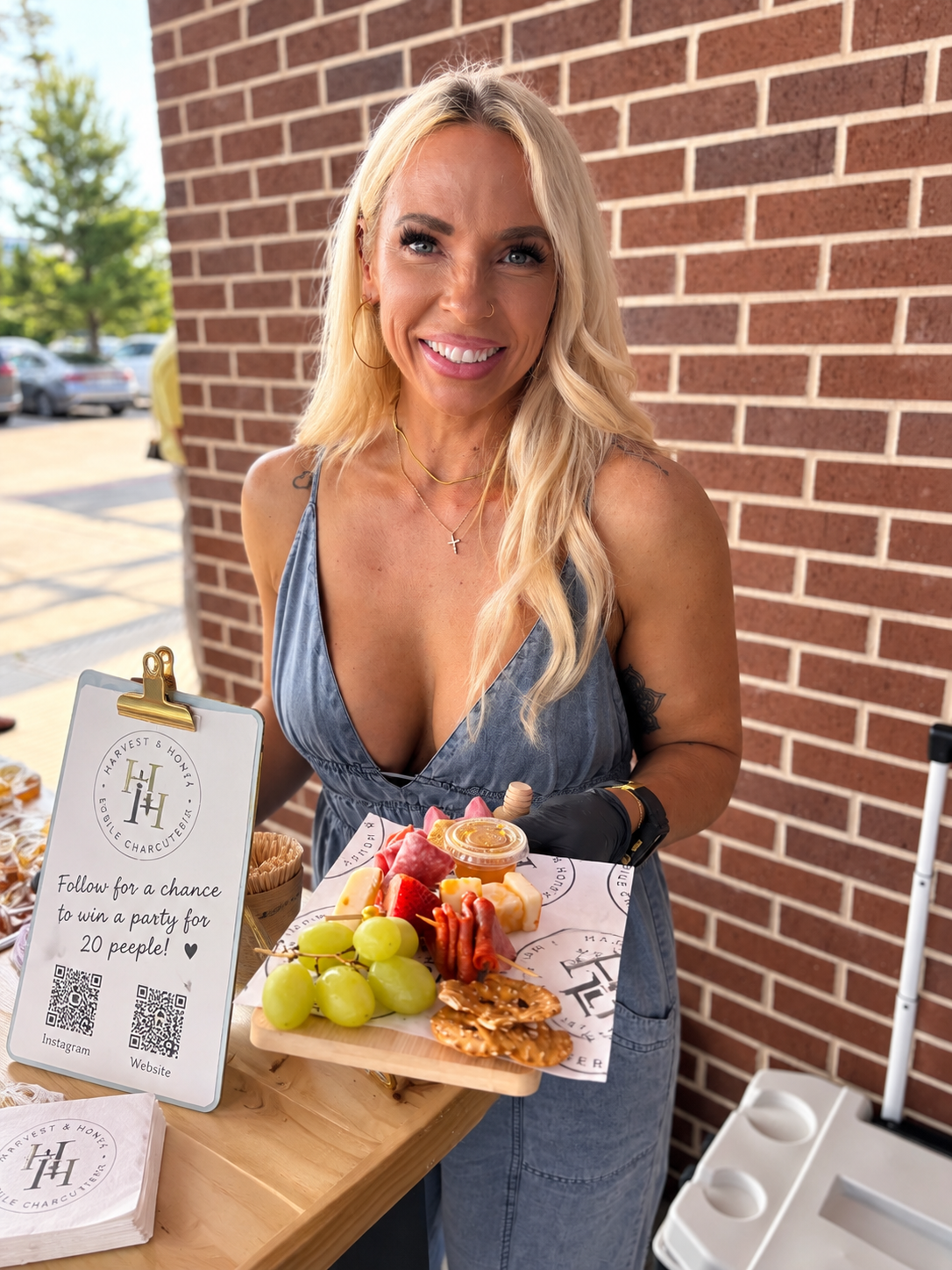 A smiling blonde woman holds a wooden platter with grapes, strawberries, cheese, and crackers at an outdoor market stall.