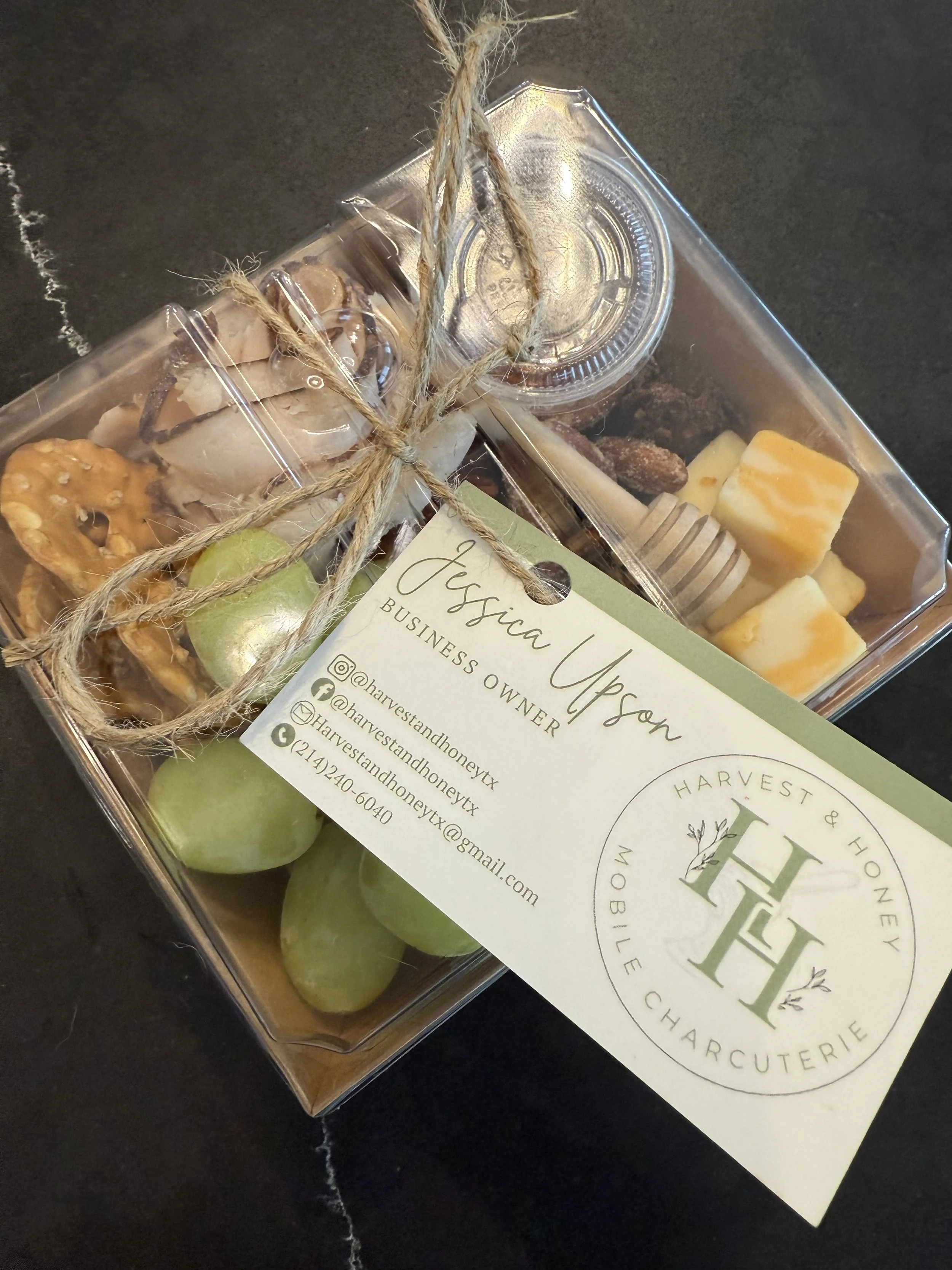 Clear plastic gift box containing green grapes, a jar of honey, assorted chocolates, and a small jar of bites, tied with twine, with a business card for harvest and honey