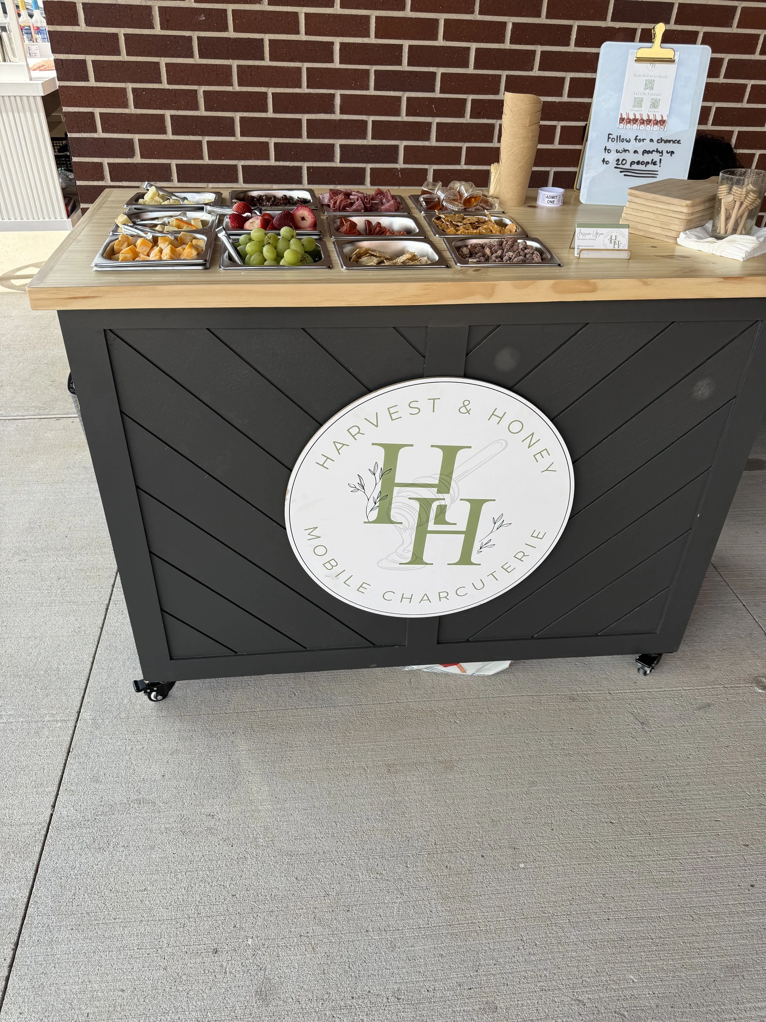 A black mobile charcuterie cart with a wooden top, featuring a white round logo with green and black text that reads 'Harvest & Honey' and 'Mobile Charcuterie,' positioned on a concrete sidewalk in front of a brick wall. The cart has wheels at the bottom. There are various containers on top of the cart filled with sliced fruit, grapes, cured meats, nuts, and other snacks. To the right of the cart, there is a sign with a QR code and text about winning a party for up to 20 people. The sign also has a rolled-up paper towel and a container holding small cups or containers.